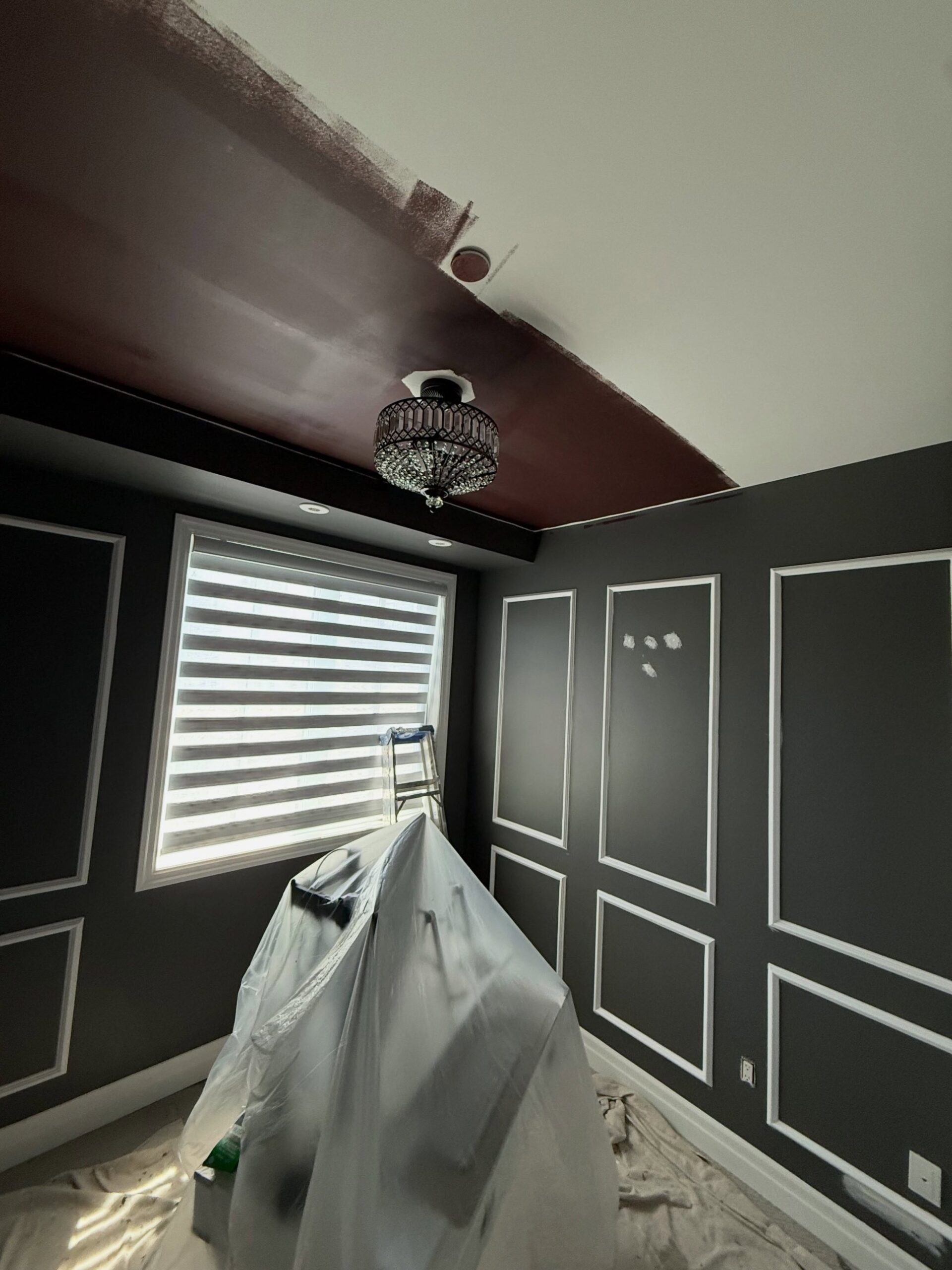 A ceiling halfway painted deep burgundy over the original white, with a crystal flush-mount chandelier at the center, two small recessed can lights, and a plastic drop cloth draped over furniture below.