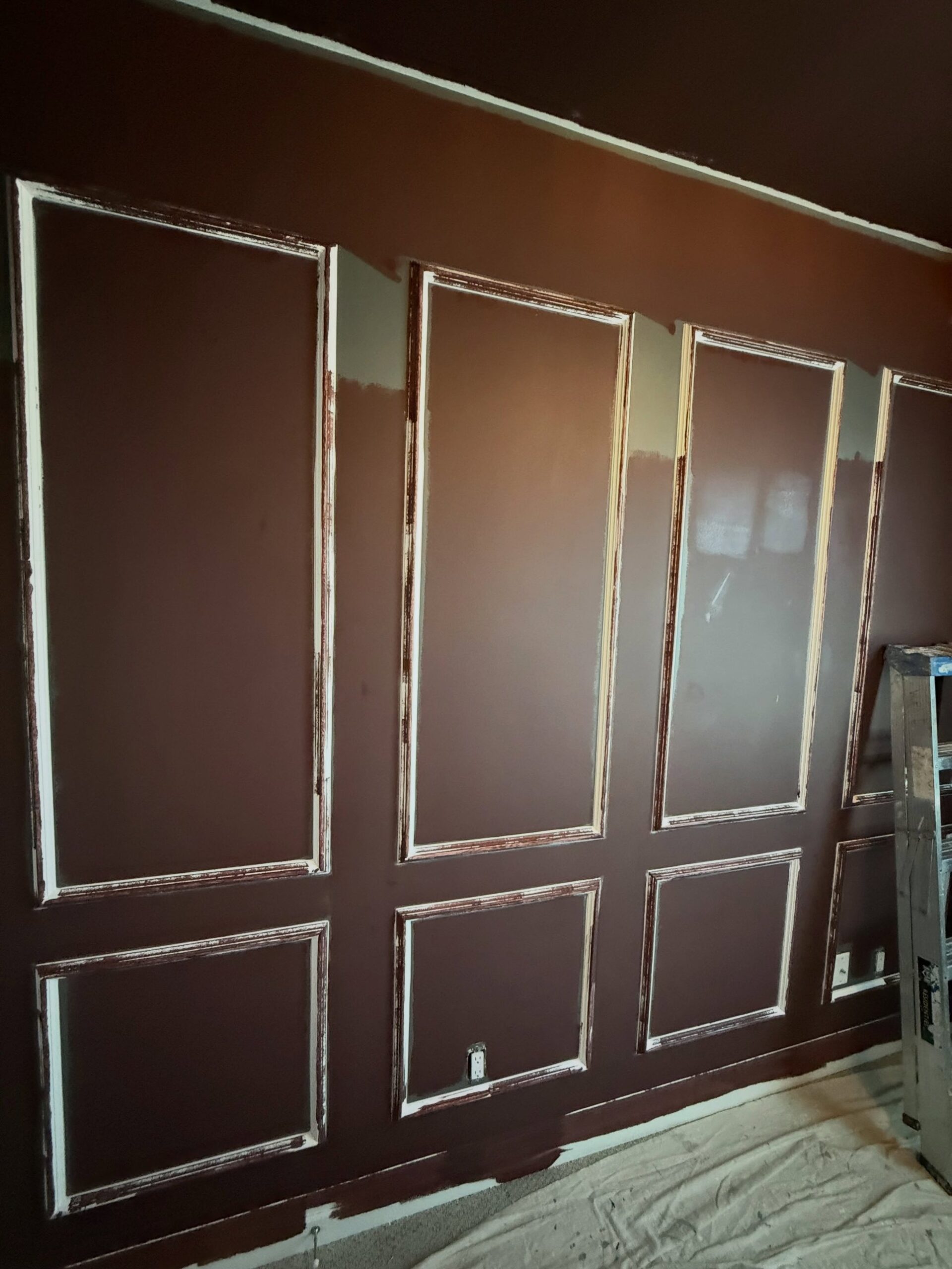 A burgundy wall with panel moldings mid-paint, white primer still peeking around the edges of each frame and lighter raw streaks showing where the burgundy hasn't fully covered the trim yet.