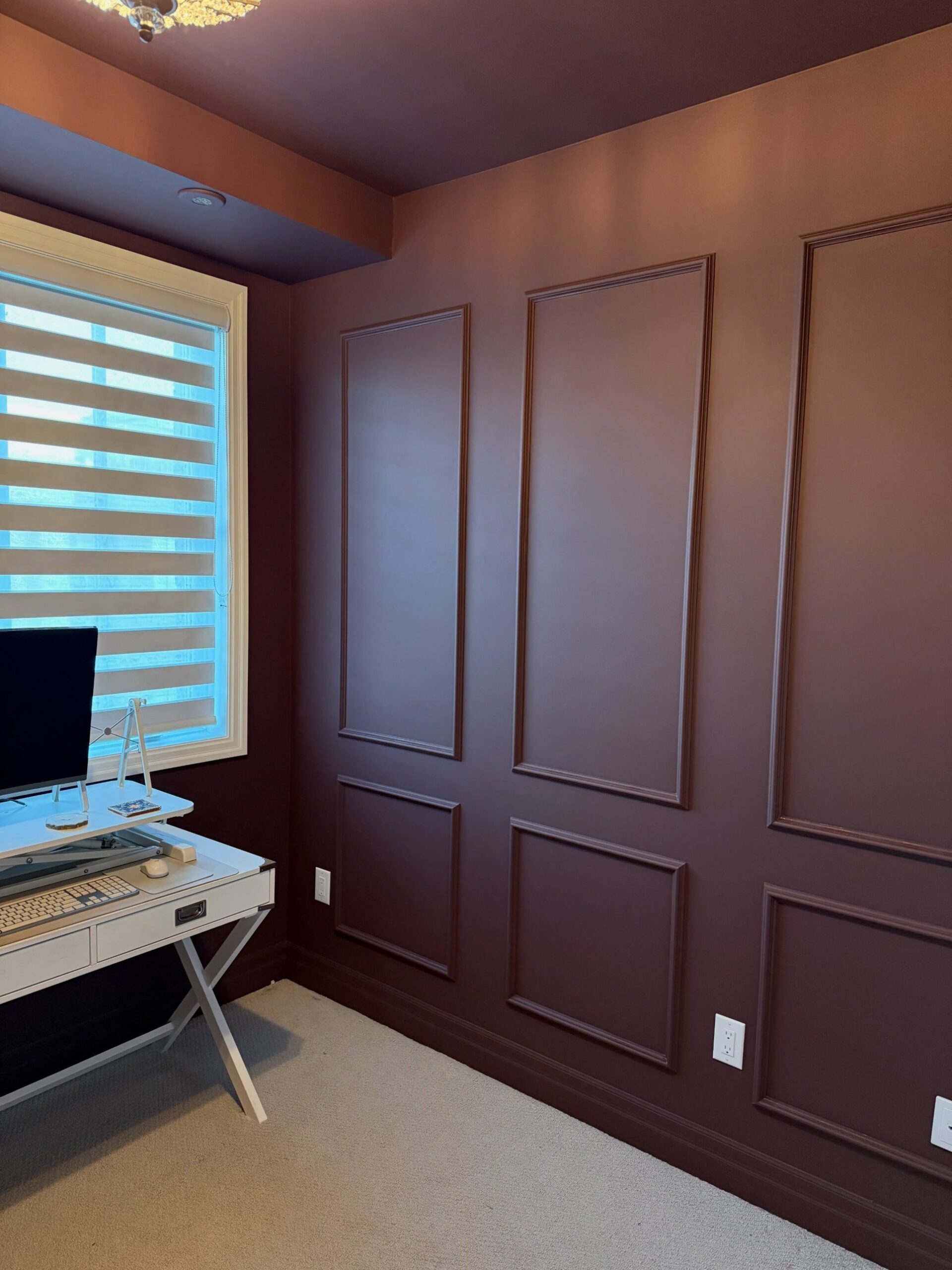 The same office in burgundy, with the white campaign desk returned under the window, a monitor on a riser, a crystal flush-mount ceiling light, and the burgundy panel wall running from window to corner.