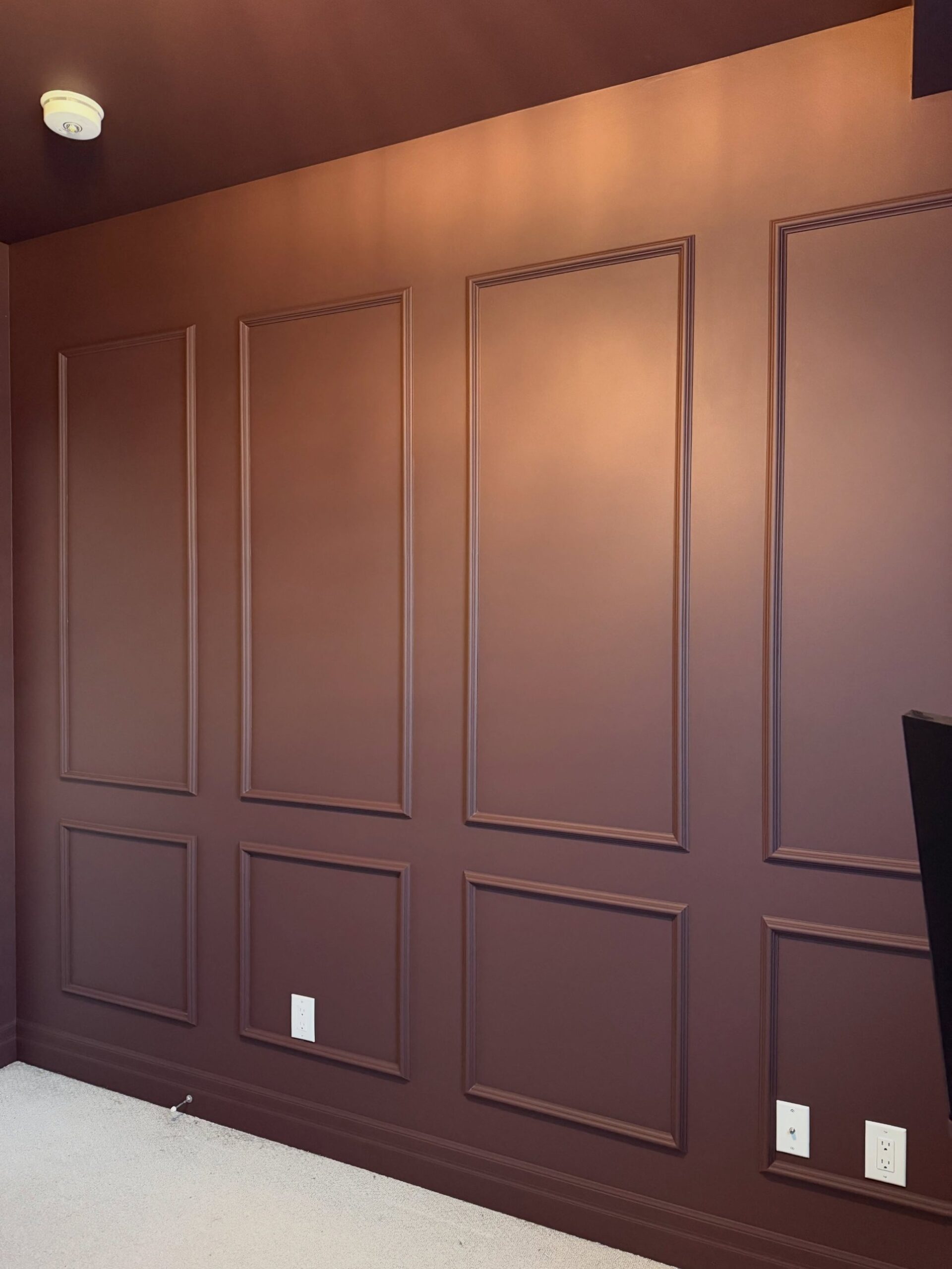 A fully drenched burgundy wall with four tall panel moldings on top, four smaller square panels below, baseboards and ceiling in the same burgundy, three white outlet covers, a cream smoke detector on the ceiling, and cream carpet below.