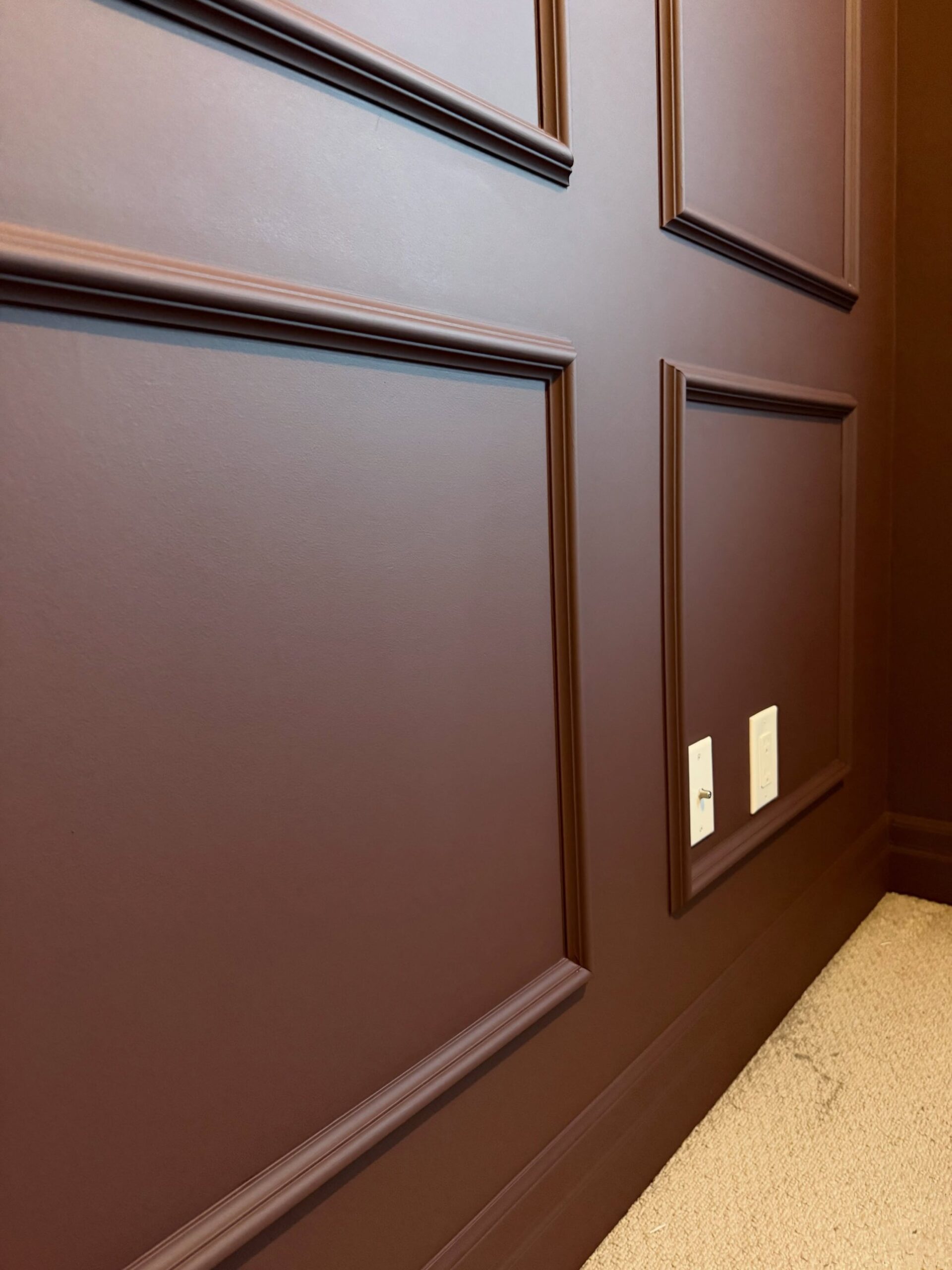 Close-up of burgundy panel moldings showing shadow lines along each trim profile, a chair-rail band, the baseboard also painted burgundy, two small white switch plates, and cream carpet below.