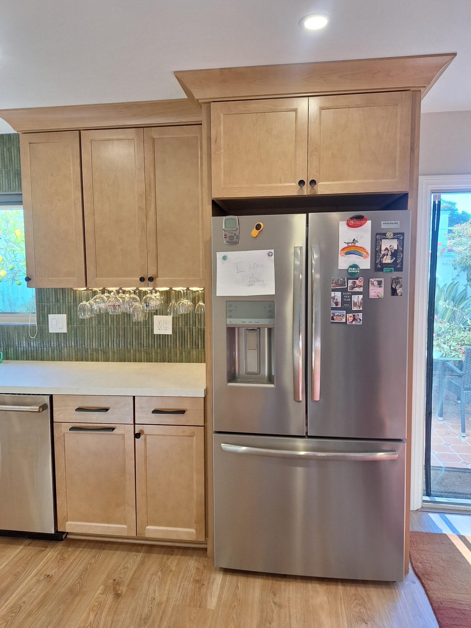 Stainless French-door fridge tucked into a cabinetry surround with a matching upper above, a wine glass rack mounted under the next cabinet, and a sliding patio door to the right.