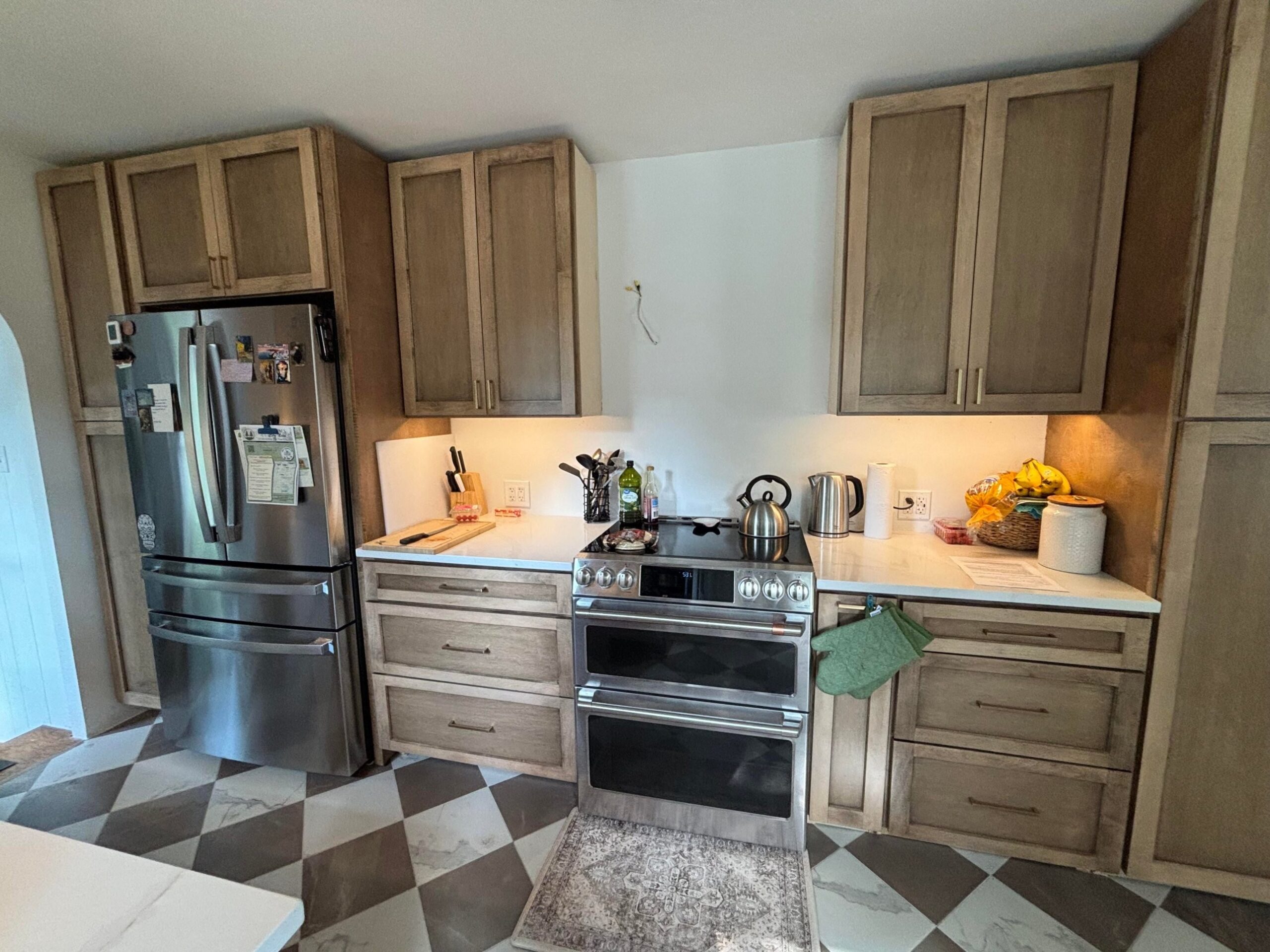 Stainless Cafe double oven induction range between stained wood cabinets, a French-door fridge on the left, light quartz counters, a kettle and an electric water boiler on the counter, and a basket of bananas and a ceramic canister on the right.