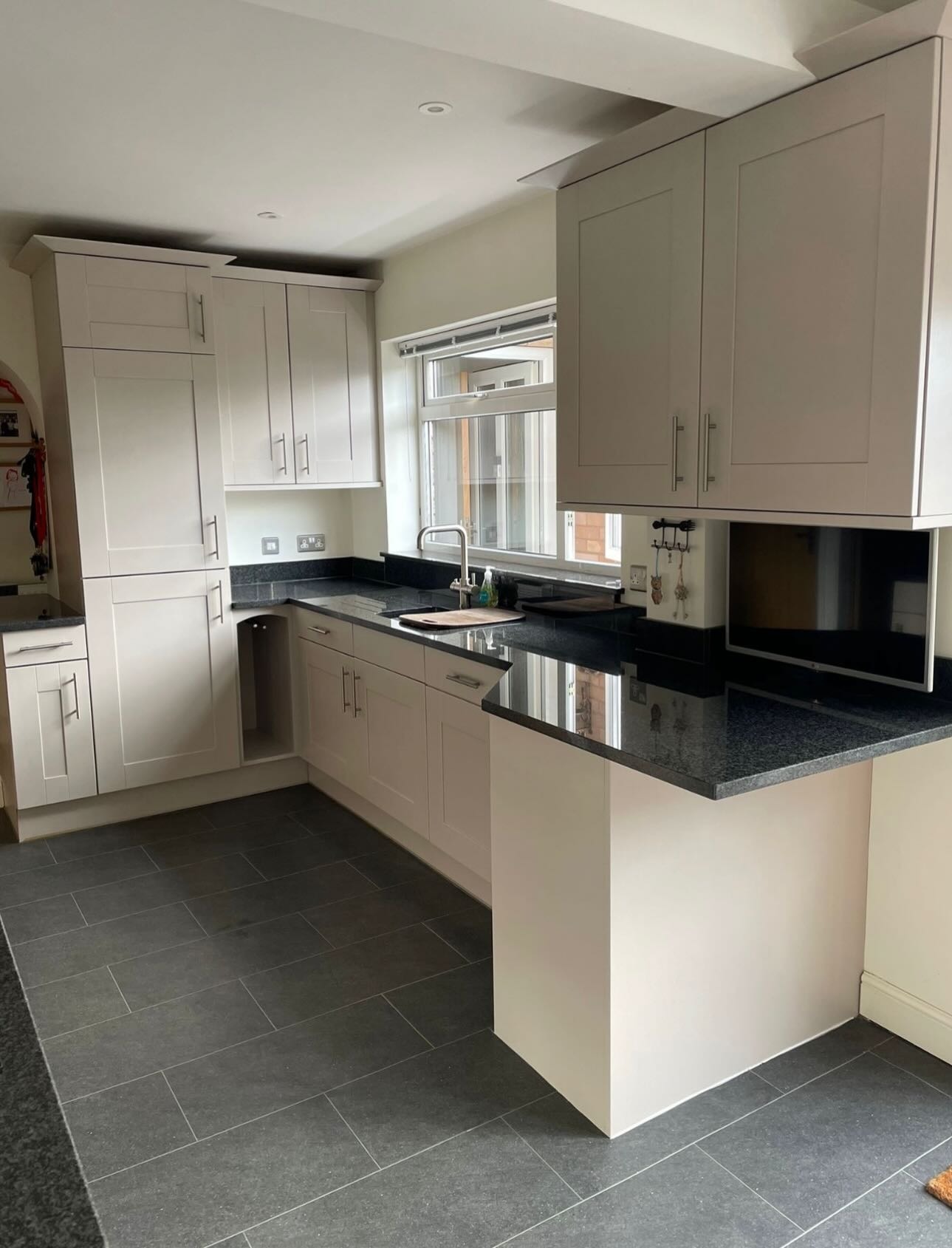 Full view of the finished cashmere kitchen with a window above the sink, tall larder cabinet in the corner, and black granite worktops throughout.
