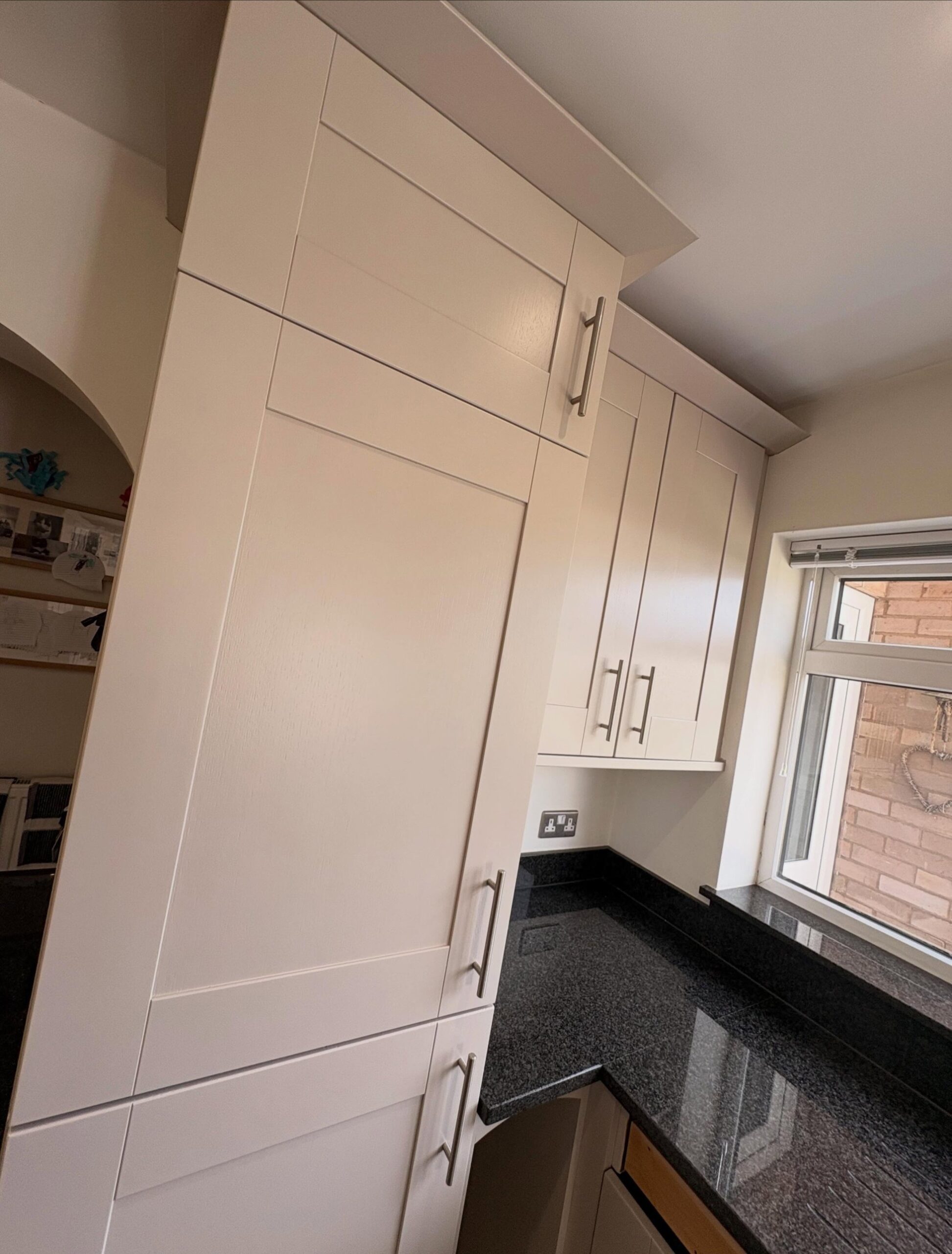 Full-height larder cabinet in cashmere satin finish beside a window, with the black granite worktop running underneath.