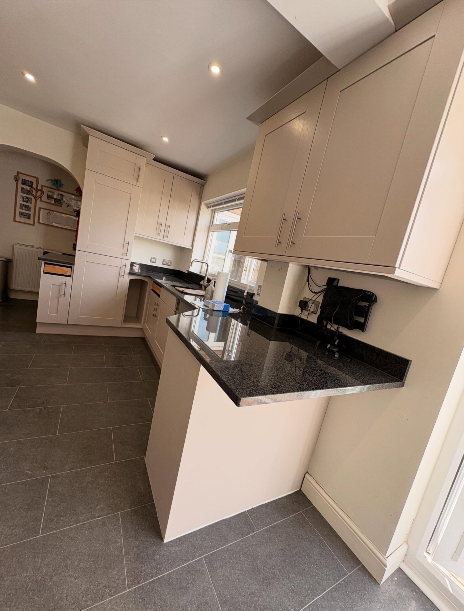 Finished U shaped kitchen in a soft cashmere greige satin paint, with black granite worktops, stainless handles, a window to the garden, and a corner microwave shelf.
