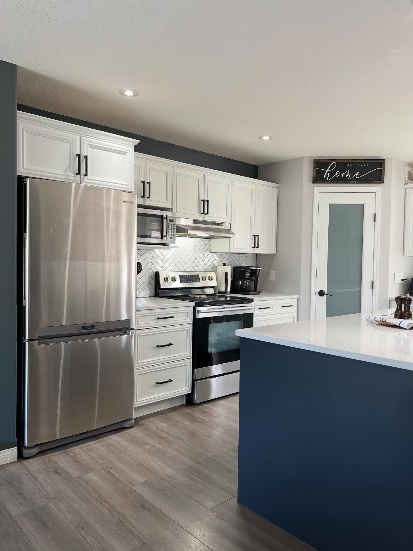 The same range wall after the cabinets were painted Chantilly Lace white with matte black bar pulls. Fridge, range, microwave, quartz counter, herringbone backsplash, and wood-look floor are all the same.