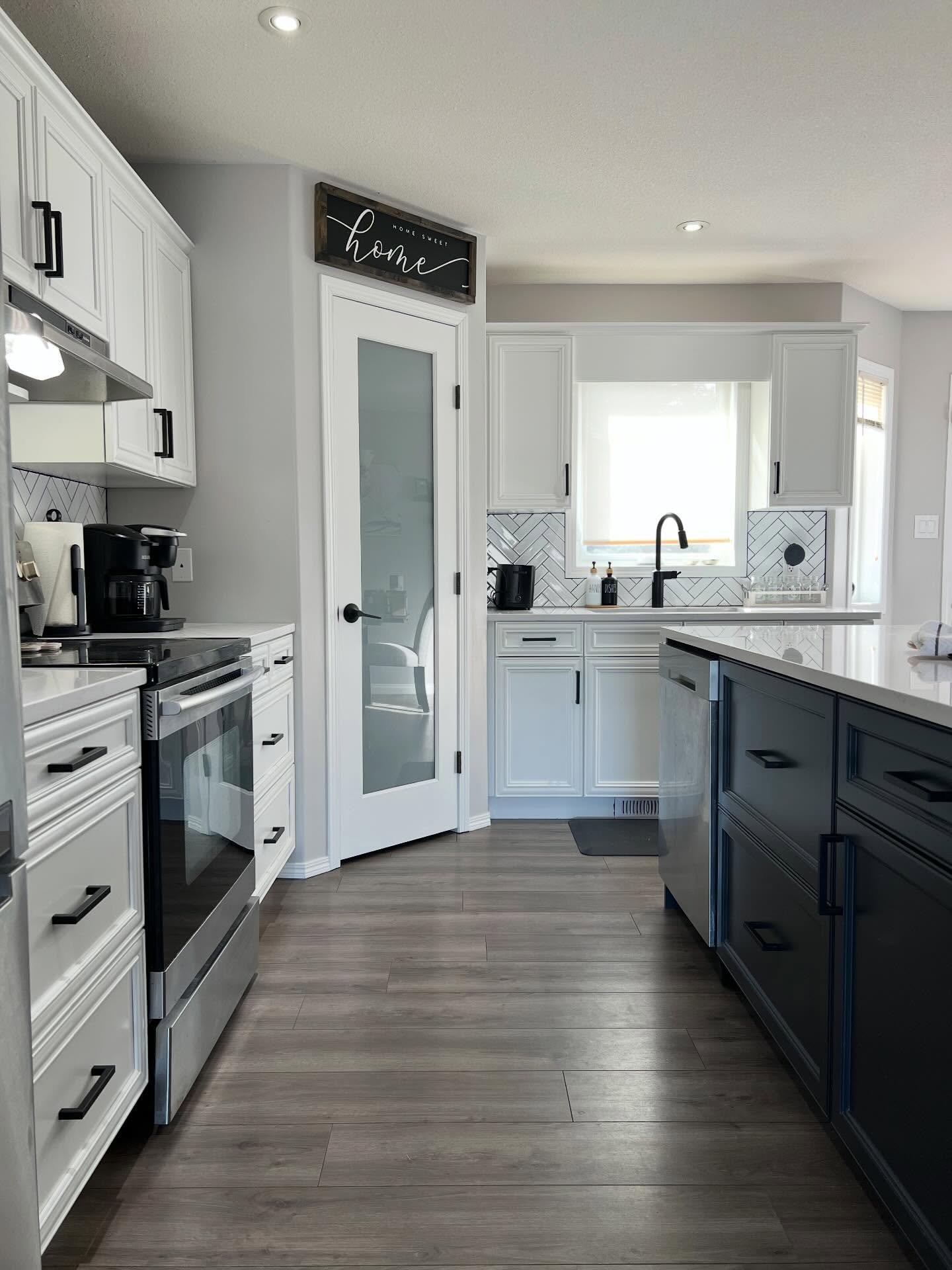 Same angle after the refresh. White painted upper and lower cabinets, a white framed frosted glass pantry door with a matte black lever handle, a black faucet above the sink window, more daylight bouncing around the room, and the navy island now reading as a clean deliberate accent.