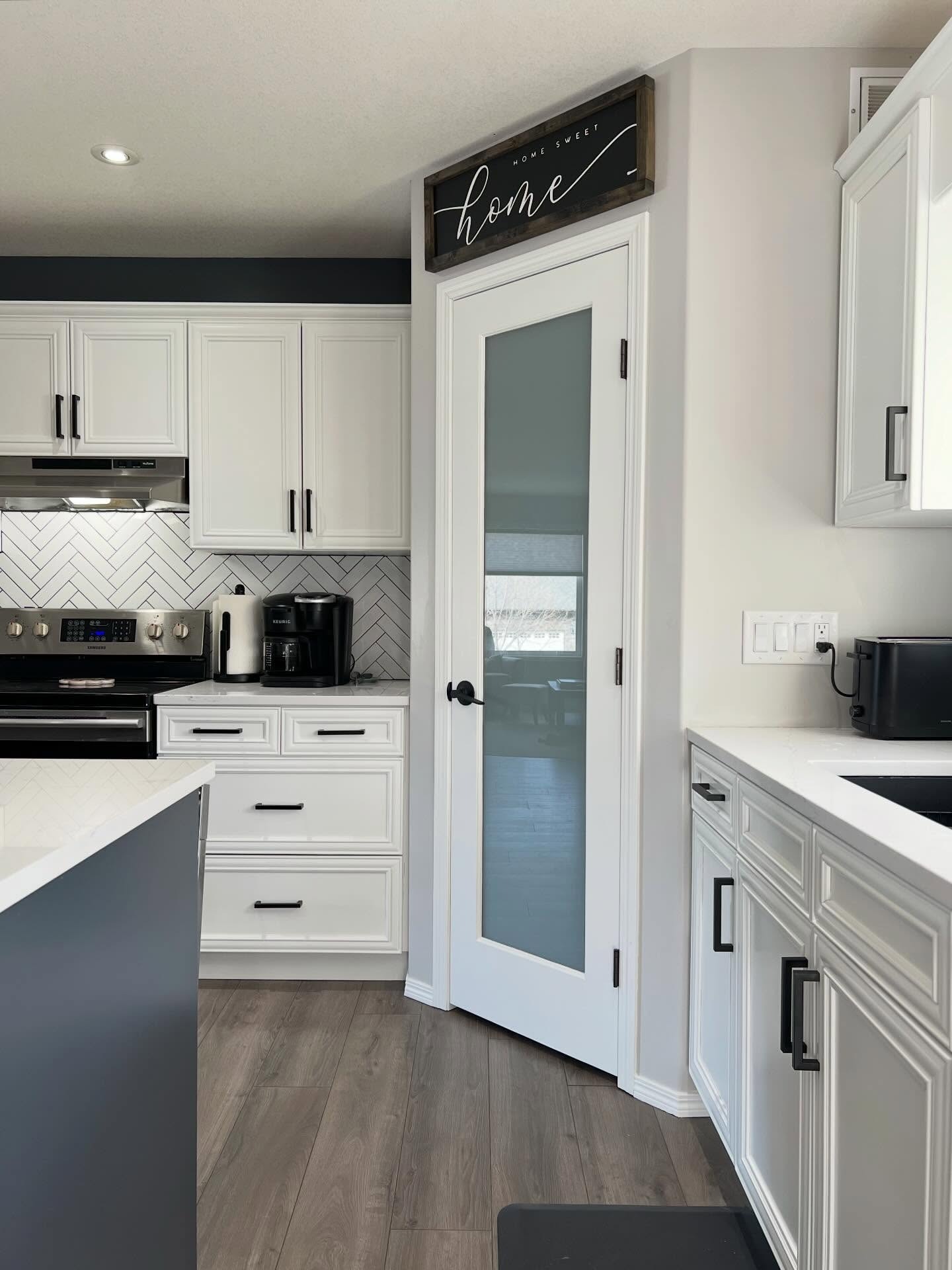Close-up of the frosted-glass pantry door painted white with a matte black lever handle, the "home sweet home" sign above it, surrounded by Chantilly Lace cabinets and matte black bar pulls.