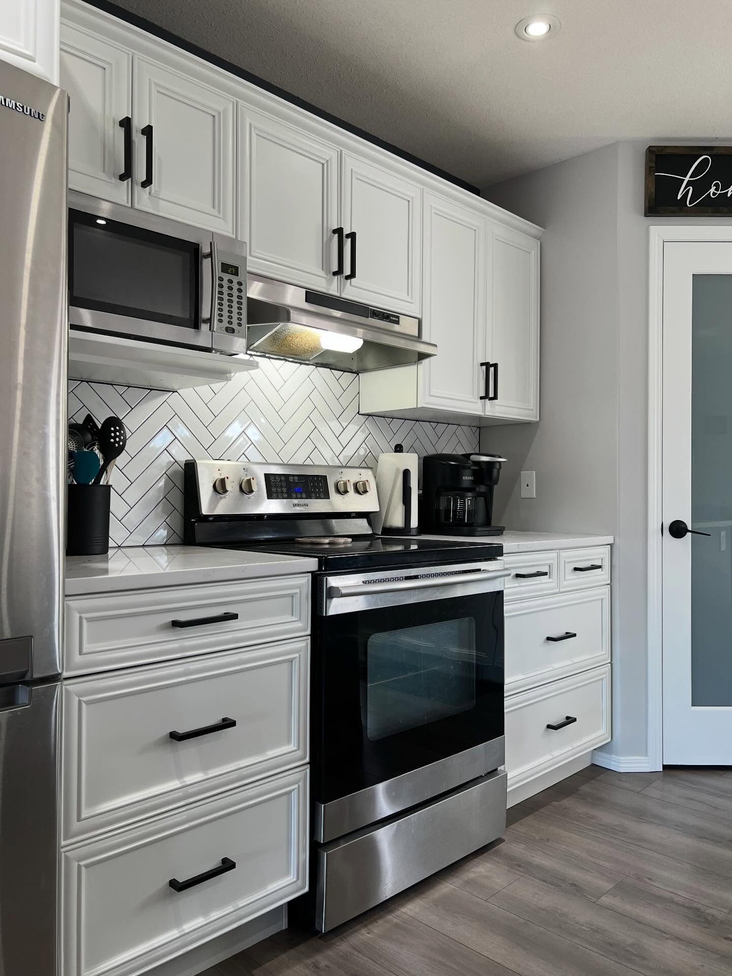Close up of white painted raised panel cabinets with squared matte black bar pulls, a Samsung stainless range, a stainless over-the-range microwave with vent hood, a black utensil crock on the quartz counter, and a bright white herringbone tile backsplash that now reads clean and modern.