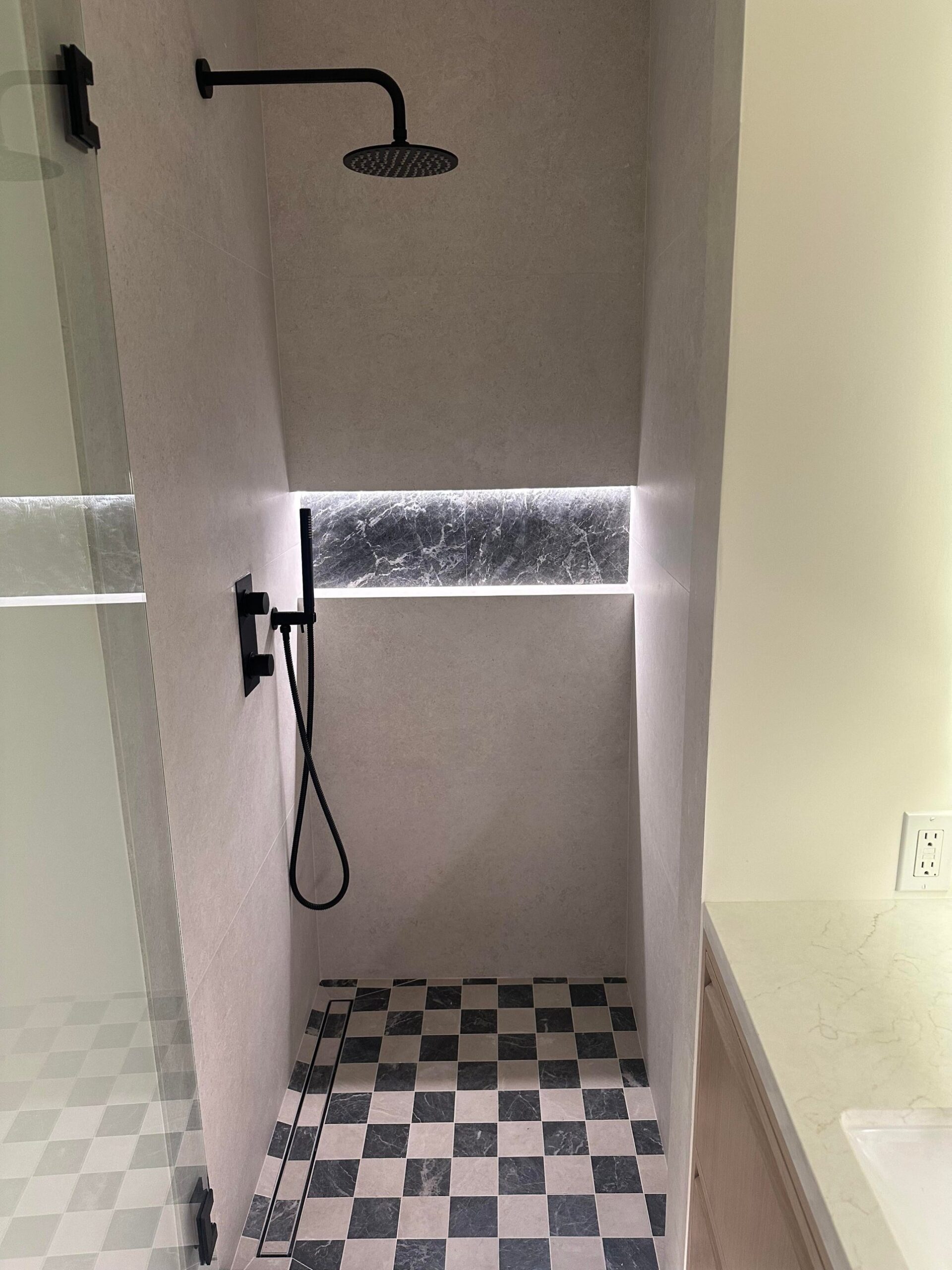 Inside the new shower with a black and white checkered marble floor, a black rain shower head, a handheld sprayer, a linear drain along the entry, and a marble shower niche with a soft LED glow at the top.