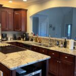 Wide landscape view of a dated cherry cabinet kitchen with a small granite island, built in wine fridge and an arched opening to a vaulted family room, shown as the before shot.
