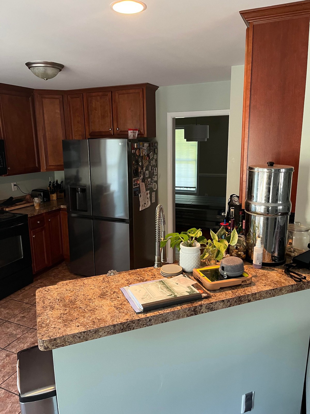 Before photo of a kitchen showing cherry-orange upper and lower cabinets, a black electric stove on the left, a black stainless LG side-by-side fridge, tan tile floor, and the kitchen peninsula with laminate counters in the foreground.