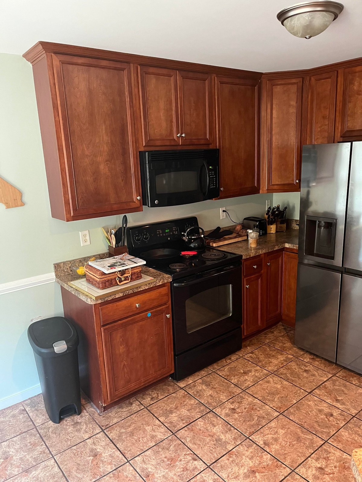 Before photo of a kitchen corner with cherry-orange raised panel upper and lower cabinets, a black electric stove, a black GE over-the-range microwave, a tan tile floor, and a sage green wall peeking in on the left.