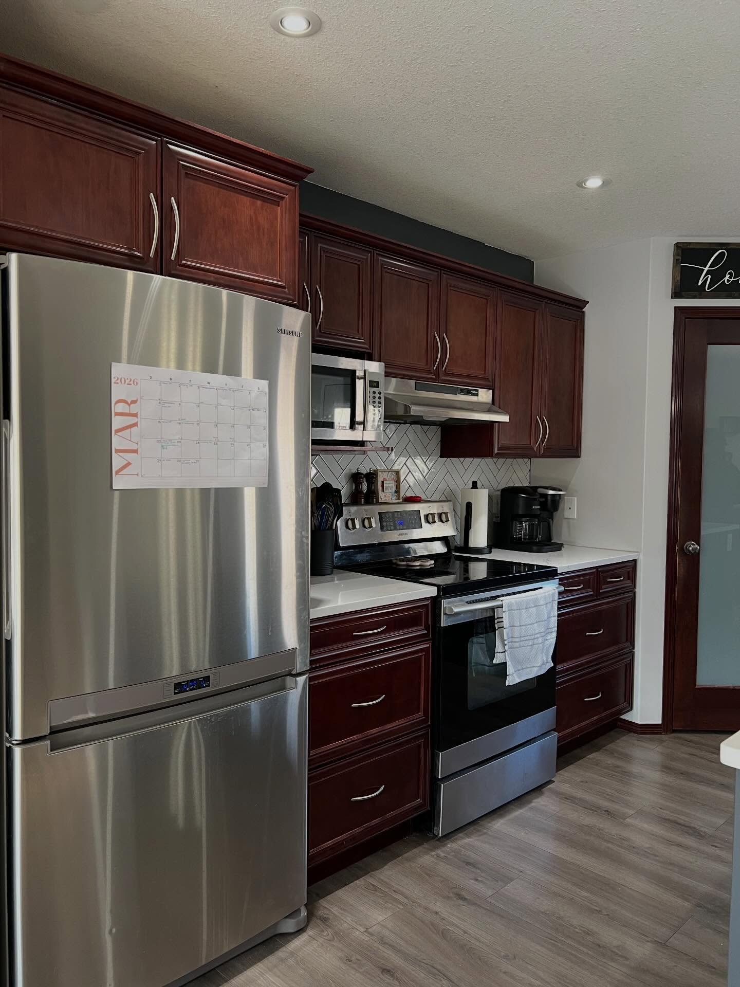 Dated kitchen with reddish brown cherry raised panel cabinets, a Samsung stainless fridge with a MAR 2026 wall calendar, a stainless range, white herringbone tile that reads yellow against the dark wood, and a frosted glass pantry door framed in cherry.