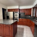 Dated kitchen before remodel with orange cherry cabinets, arched cathedral cabinet doors, speckled green granite countertops, double wall oven next to stainless fridge, white tile floor, and matching cherry wood island.