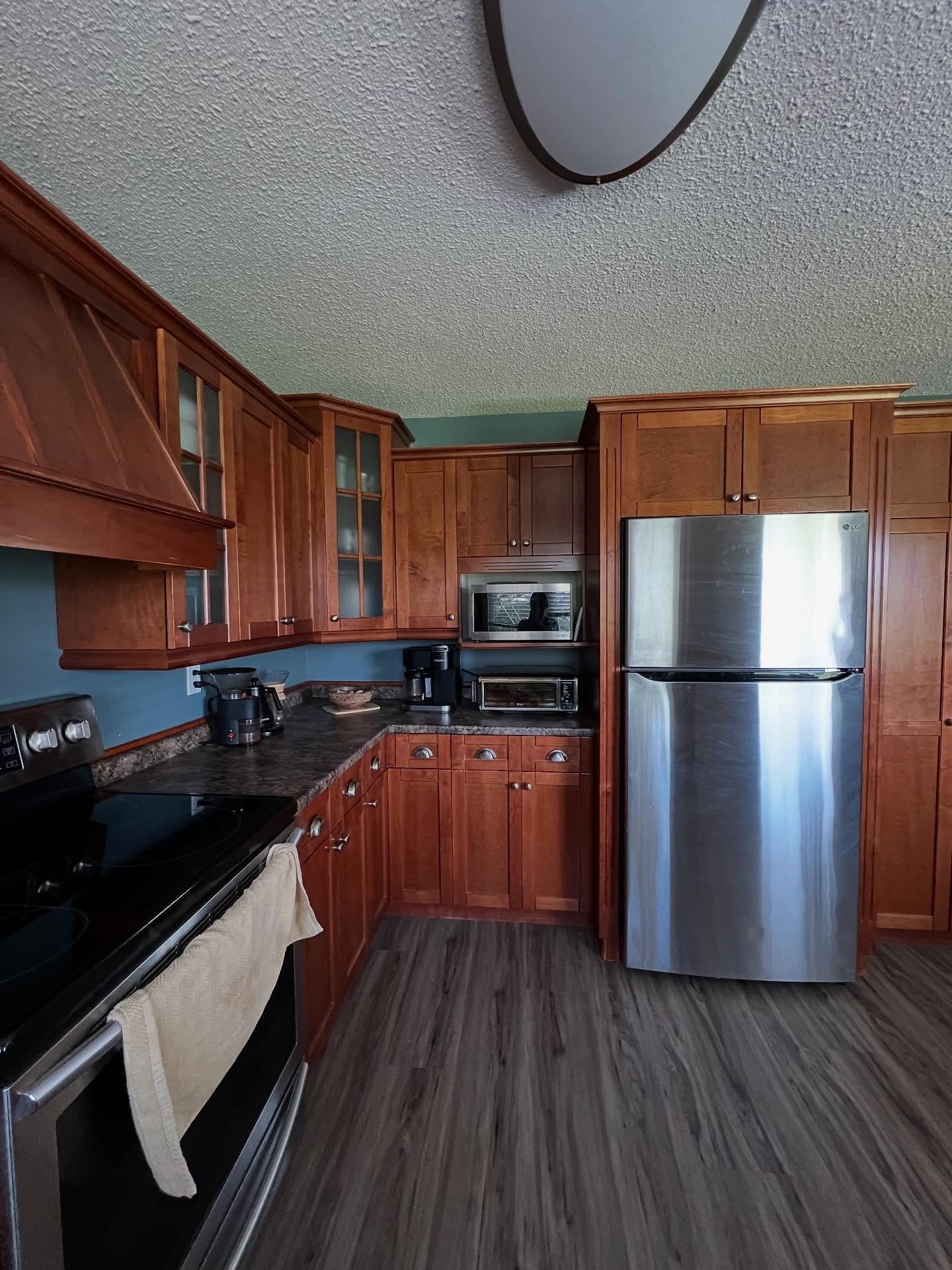 Corner view of a cherry wood kitchen showing a stainless steel top-freezer fridge, a built-in microwave, a toaster oven, teal walls, popcorn ceiling, and gray plank flooring.