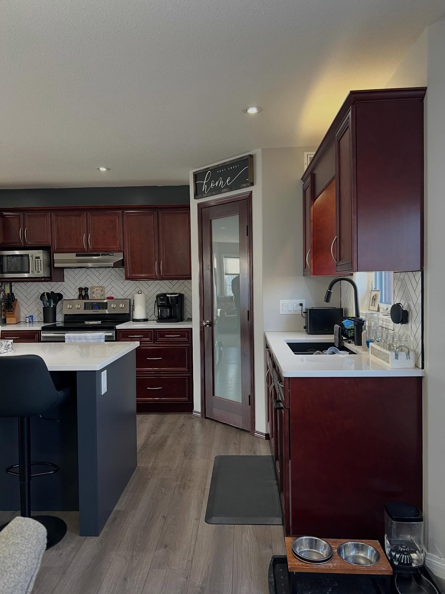 Side view of the cherry kitchen showing the navy painted island with a black bar stool on the left, cherry sink cabinets on the right with a black faucet, a cherry pantry door in the middle, and a dog feeding station on the floor near the sink.