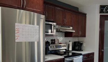 Dated U-shaped kitchen with reddish brown cherry raised panel cabinets, a Samsung stainless fridge with a MAR 2026 wall calendar taped to the door, a stainless range with over-the-range microwave, white herringbone subway tile backsplash reading yellow against the dark wood, charcoal paint band above the uppers, and a frosted glass pantry door framed in cherry.