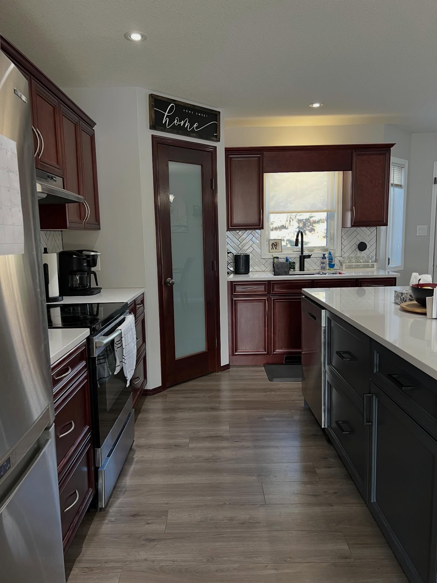 Pantry corner view in the cherry kitchen showing a cherry framed pantry door with a frosted-glass panel, cherry upper cabinets around the window, a navy island with white quartz waterfall top, a black gooseneck faucet, and wood-look flooring.