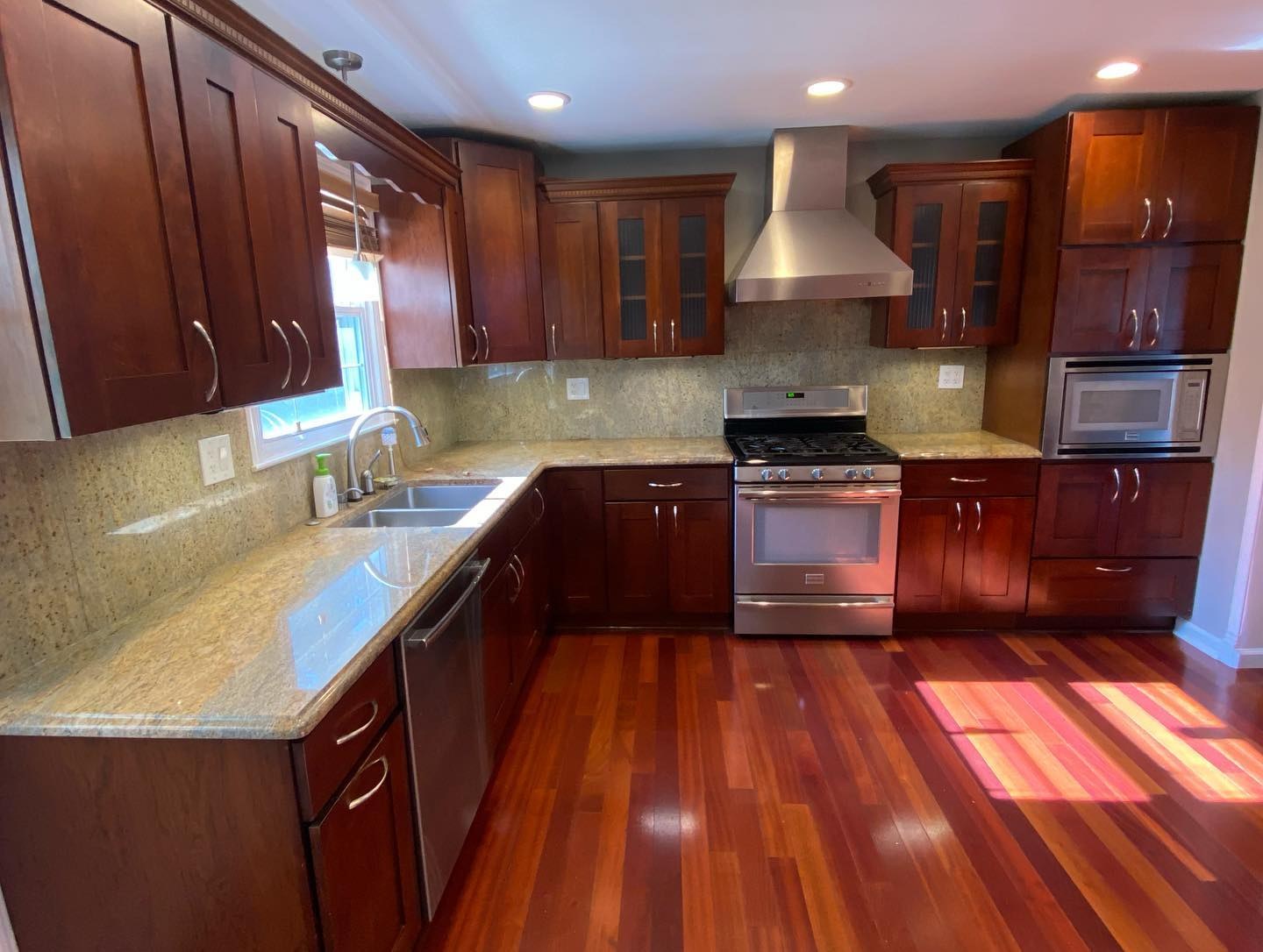 Dark cherry wood kitchen cabinets along the sink wall with granite counters, stainless hood, and glossy red Brazilian cherry hardwood floors reflecting warm light.