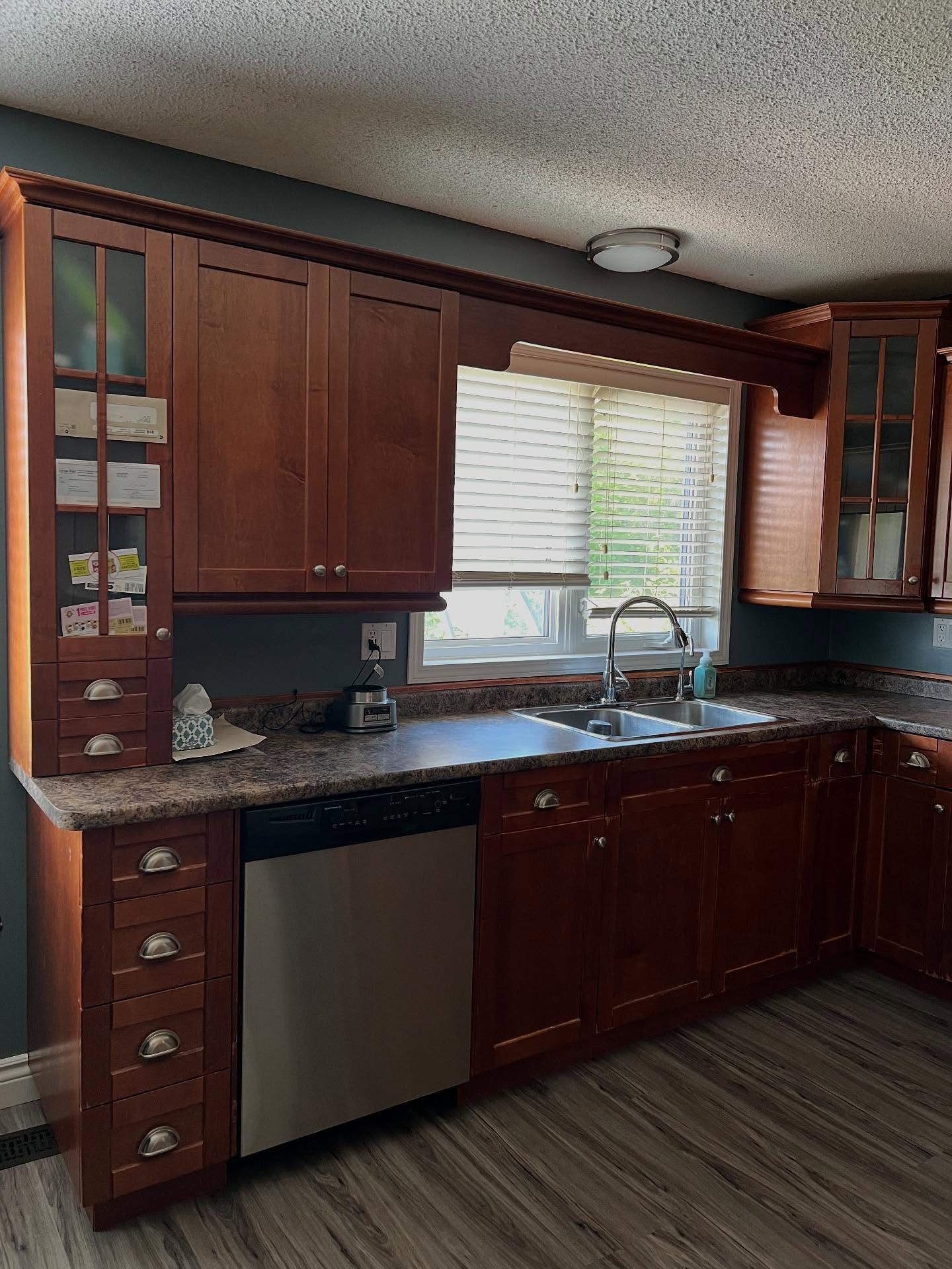 Close-up of the sink wall in a cherry wood kitchen with dark laminate counter, a stainless double-bowl sink, a window with decorative wood scrolls on either side, white blinds, and a popcorn ceiling.