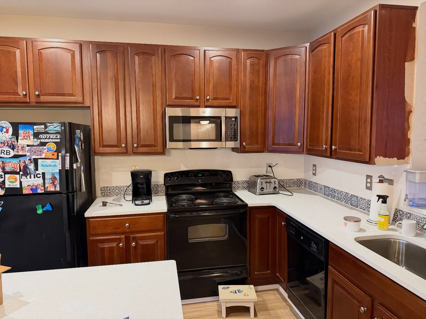 Close-up of a kitchen with cherry wood cabinets, a black electric coil range, a stainless microwave, white quartz counters, and a navy and gray Moroccan patterned tile strip running across the white subway backsplash.