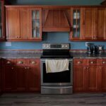 Wide landscape view of a dated U-shaped kitchen with dark cherry Shaker cabinets, a matching wood vent hood centered above a stainless steel electric range, teal painted walls, dark brown speckled laminate counters, and gray wood-look vinyl plank flooring.