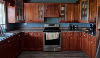 Wide landscape view of a dated U-shaped kitchen with dark cherry Shaker cabinets, a matching wood vent hood centered above a stainless steel electric range, teal painted walls, dark brown speckled laminate counters, and gray wood-look vinyl plank flooring.