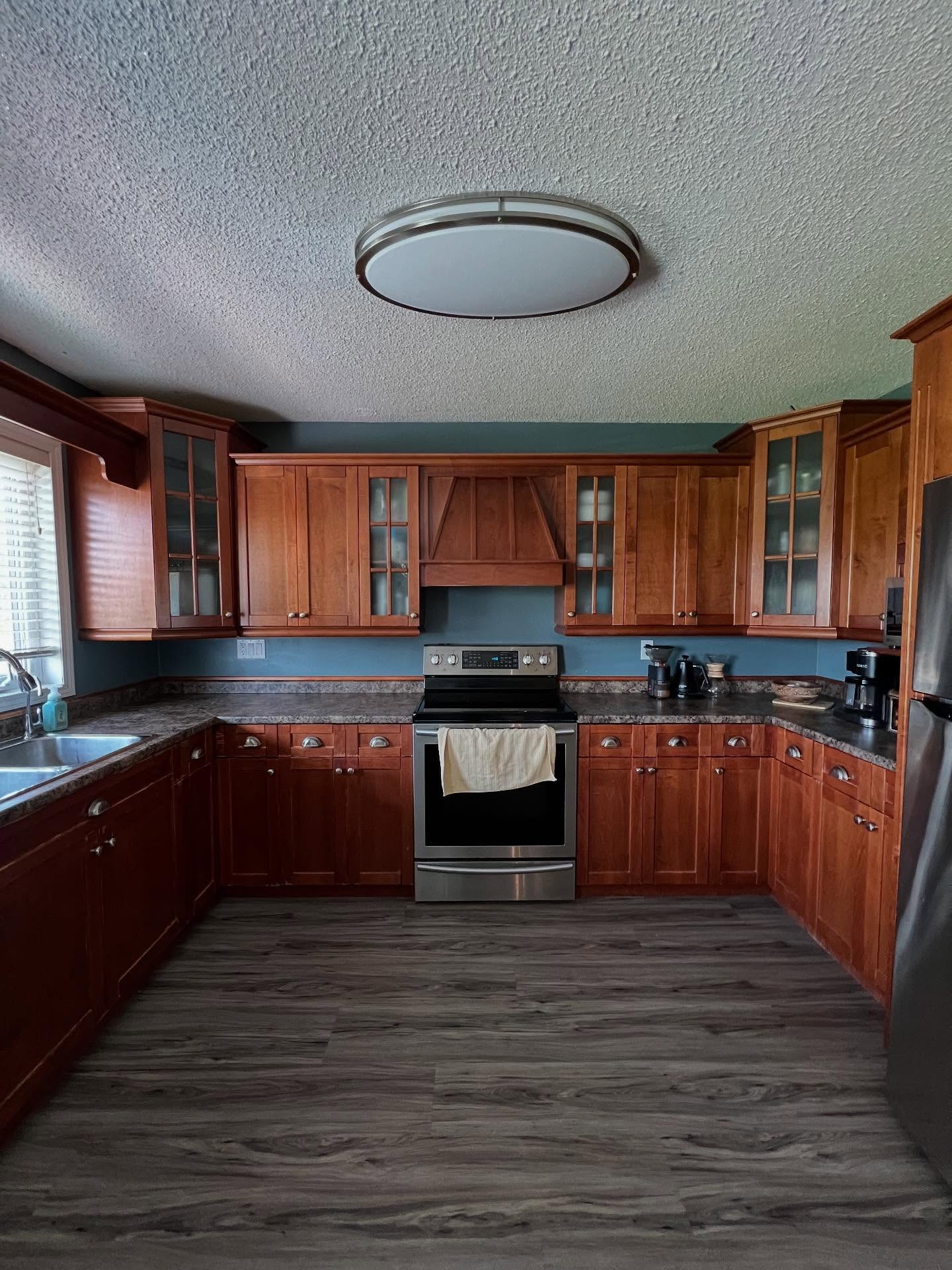Wide view of a U-shaped kitchen with dark cherry Shaker cabinets, teal painted walls, a popcorn ceiling with a large chrome dome light, dark laminate counters, a stainless steel electric range, and gray vinyl plank flooring.