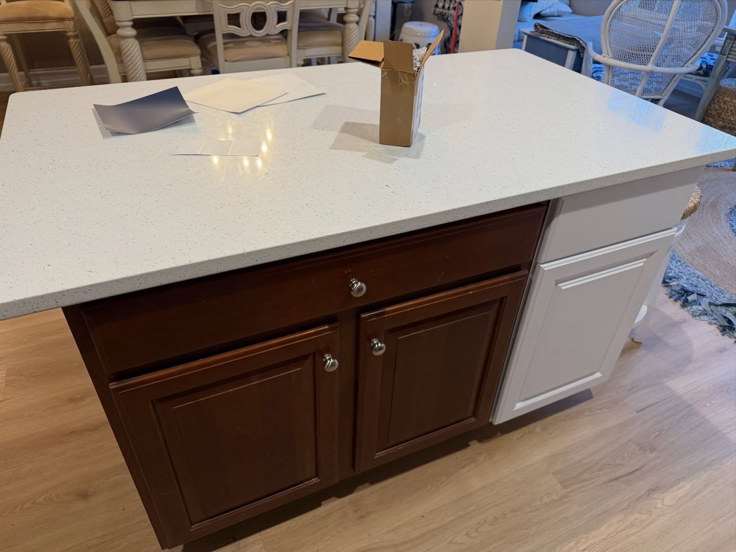 Close-up of a kitchen island with cherry wood doors and a white quartz top, with a freshly painted white cabinet visible on the right side, papers and a small cardboard box resting on the counter, and light oak flooring below.