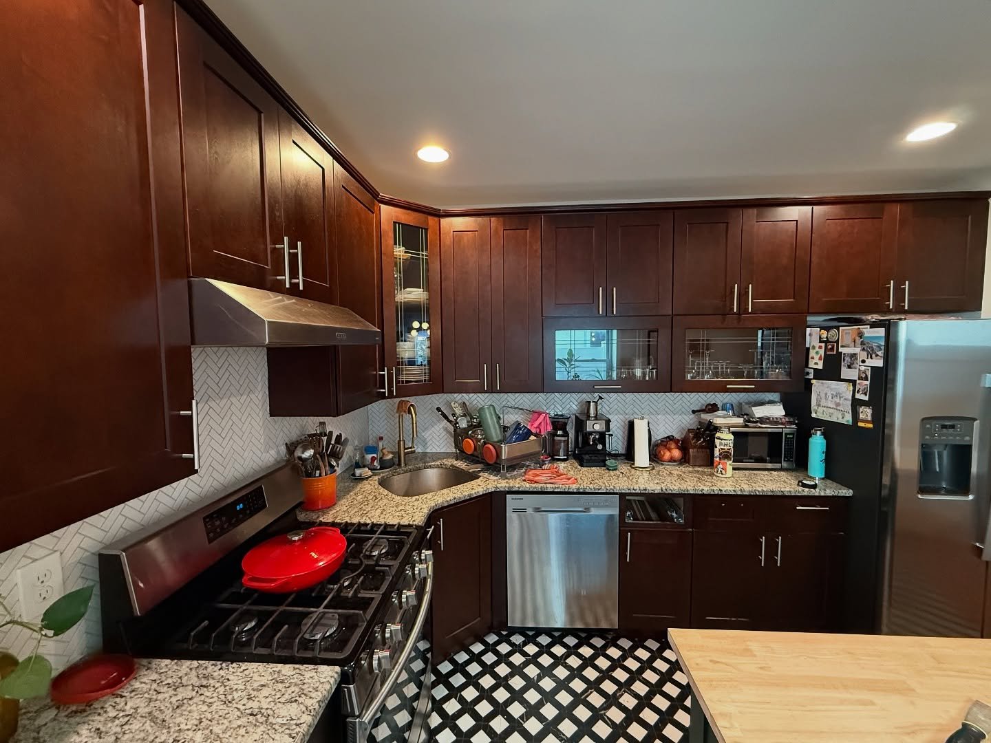 Dark cherry shaker kitchen with speckled granite counters, white herringbone backsplash, stainless hood, red Dutch oven on a gas range, and black and white checkered tile floor.