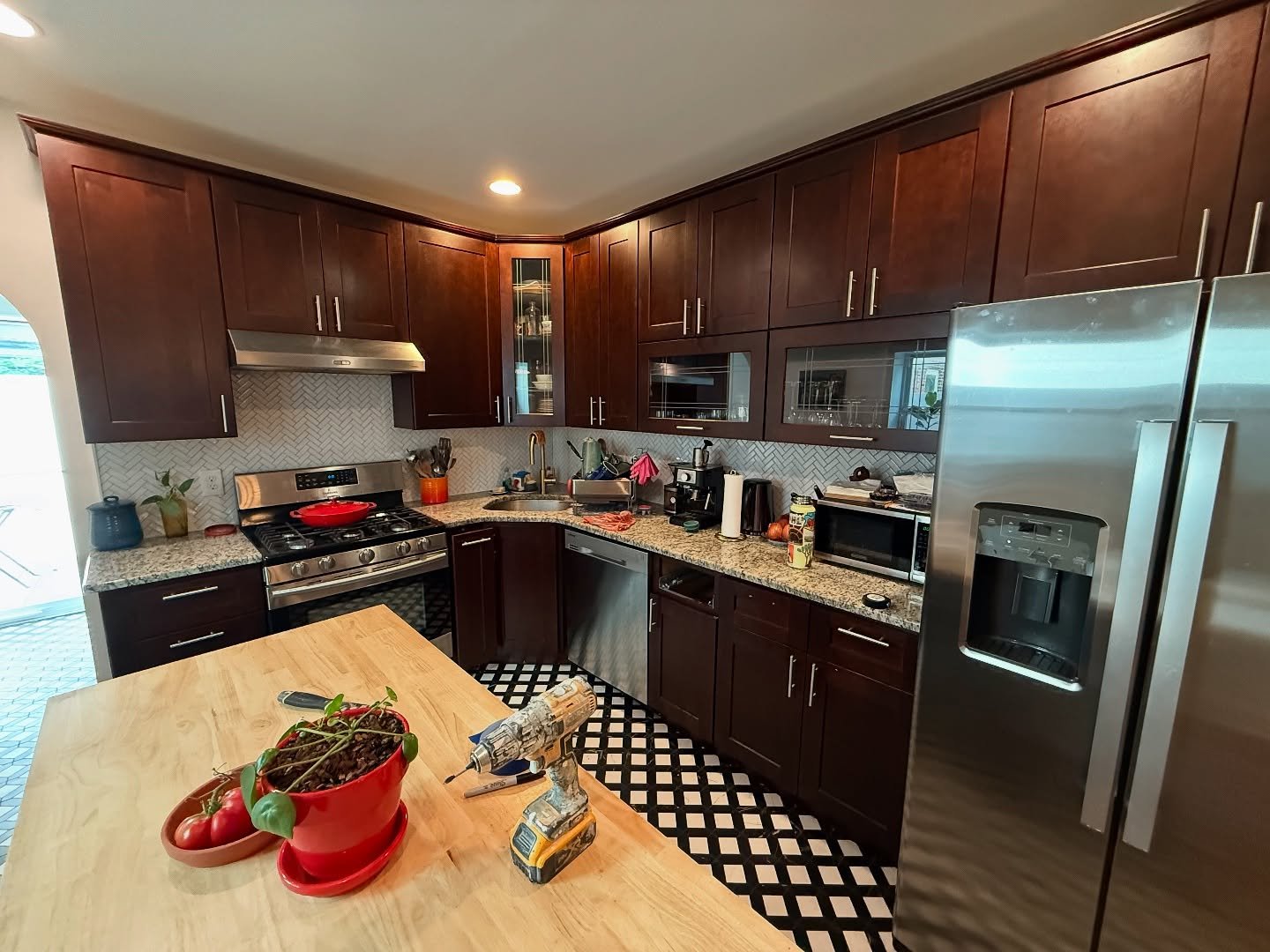 Wide view of dark cherry kitchen with butcher block island, stainless side by side refrigerator, speckled granite counters, and bold black and white checkered floor tile.