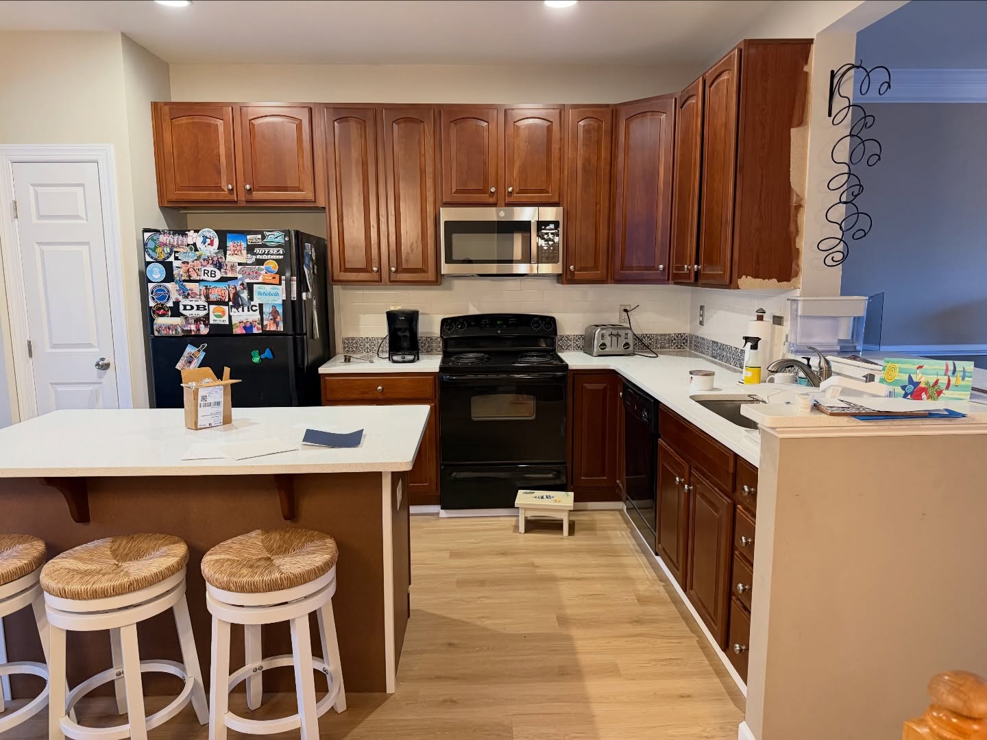Kitchen with cherry wood perimeter cabinets, a brown-paneled island base topped with white quartz, three white round rush-seat stools, a black fridge, a black stove, a stainless microwave, and light oak flooring.