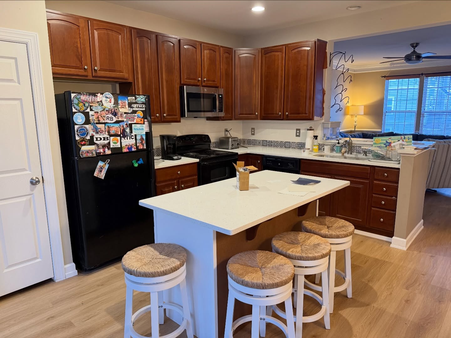 Wide view of a kitchen with orange cherry wood cabinets, a large white quartz island with four round rush-seat stools, a black fridge covered in travel magnets, a stainless microwave, a black stove, and light oak LVP floors.