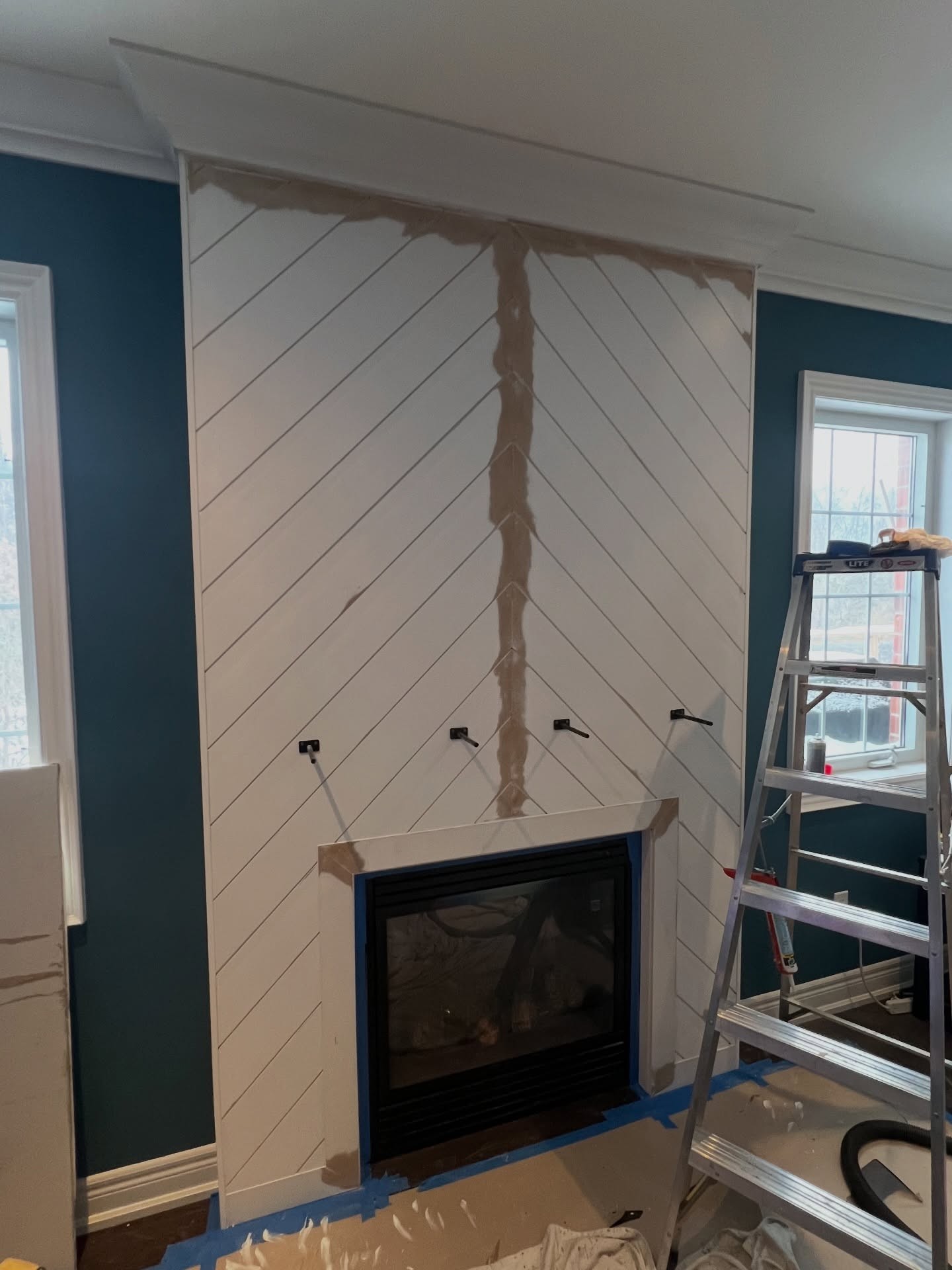 Four black iron mantel brackets sticking out from the painted chevron wall above the fireplace, ready to receive the reclaimed wood beam.