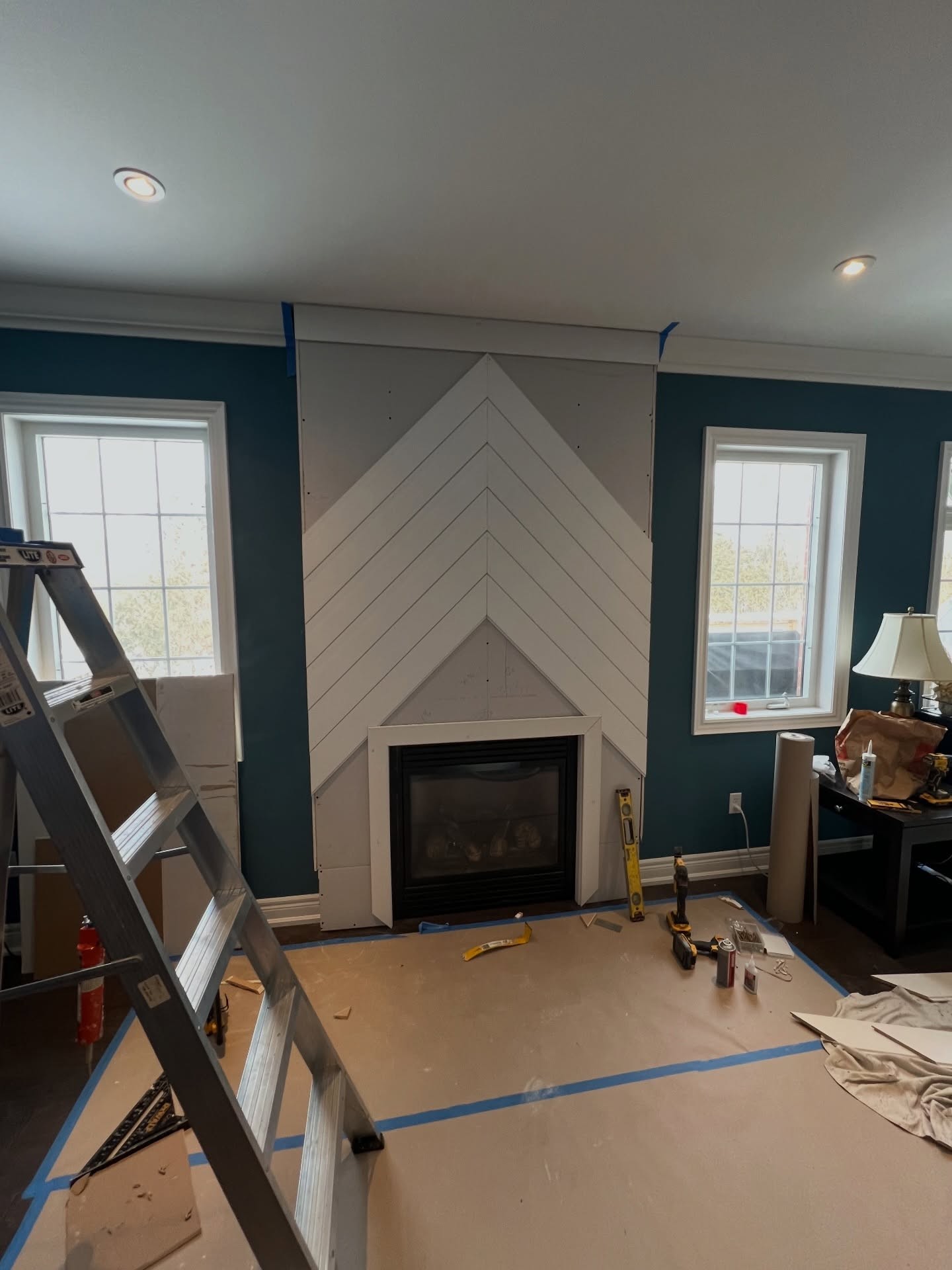 Angled view of the chevron wall taking shape between the two windows, surrounded by construction paper, ladder, and tools, with the dark teal walls still visible on each side.