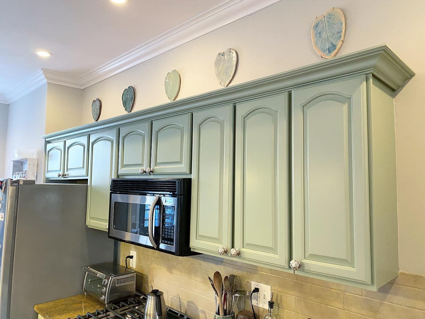 Angled view of painted sage green upper cabinets with cathedral arch doors, white crown molding above, floral ceramic knobs, and blue-painted ceramic heart decorations resting on top of the cabinets.