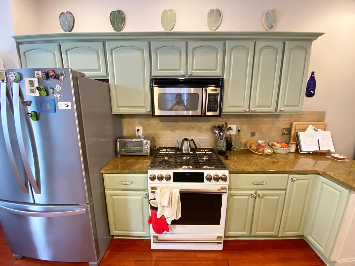 The same range wall after being painted in Sherwin-Williams Clary Sage, with soft green upper cabinets, ceramic heart decor along the top, and the original gold granite counters still in place.