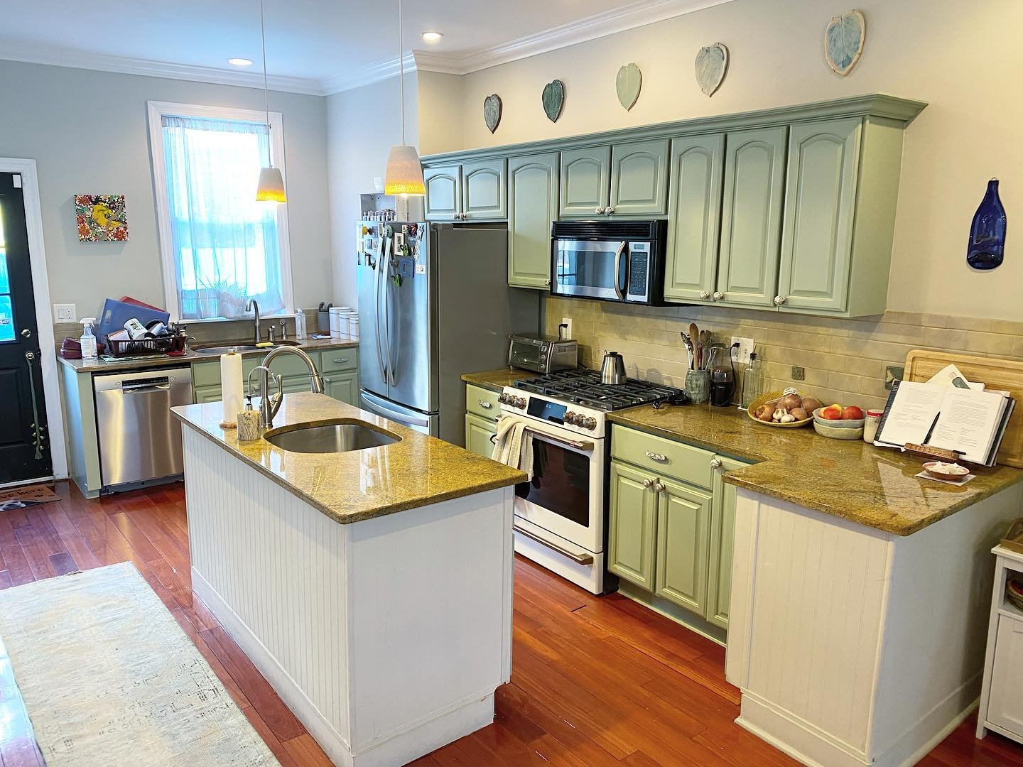 Finished kitchen view with sage green painted cabinets, white island, gold granite counters, stainless appliances, cherry hardwood floors, and ceramic heart artwork displayed on top of the upper cabinets.