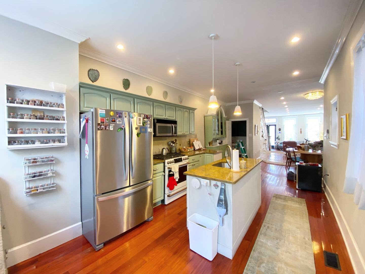 Wide hallway view of the completed Clary Sage kitchen with painted sage green cabinets, stainless French door refrigerator, white range, a small white island with gold granite top, and cherry floors.