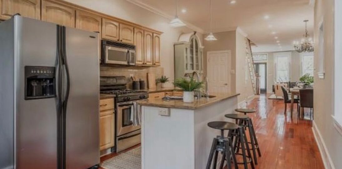 Wide view of a dated honey oak kitchen before painting, with cathedral arch upper cabinets, matching oak lower cabinets, gold speckled granite counters, a stainless side-by-side refrigerator, pendant lights over a white island with granite top, and warm cherry hardwood floors stretching down a galley layout.