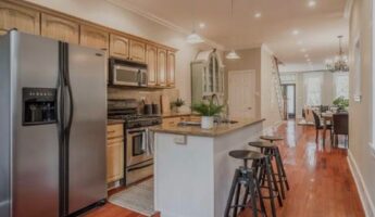 Wide view of a dated honey oak kitchen before painting, with cathedral arch upper cabinets, matching oak lower cabinets, gold speckled granite counters, a stainless side-by-side refrigerator, pendant lights over a white island with granite top, and warm cherry hardwood floors stretching down a galley layout.