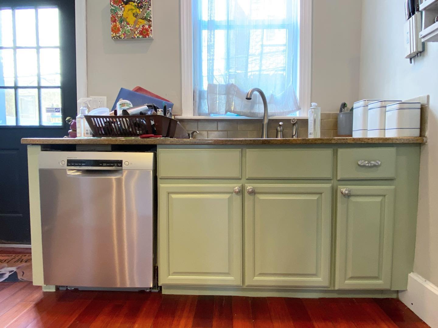 Lower kitchen cabinets painted in Sherwin-Williams Clary Sage with raised panel doors, floral ceramic knobs, stainless dishwasher, and a window above the sink with white sheer curtain.