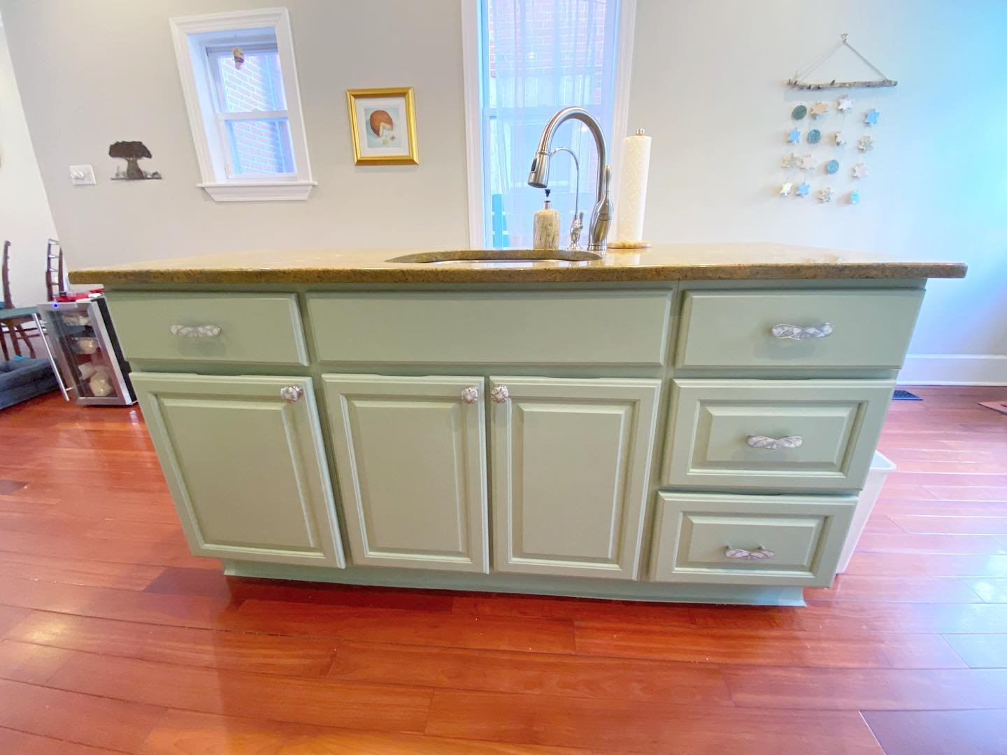 Painted kitchen island in Sherwin-Williams Clary Sage with gold granite top, floral ceramic knobs on drawers and cabinet doors, standing on warm cherry hardwood floors.