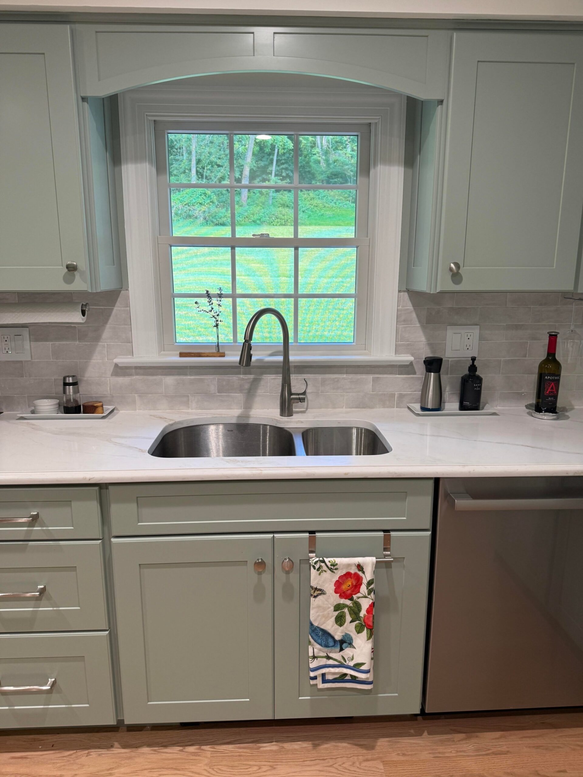Sage green shaker kitchen cabinets around a window with a soft arched valance, white quartz counter, pale gray brick tile backsplash, and a stainless pull-down faucet.