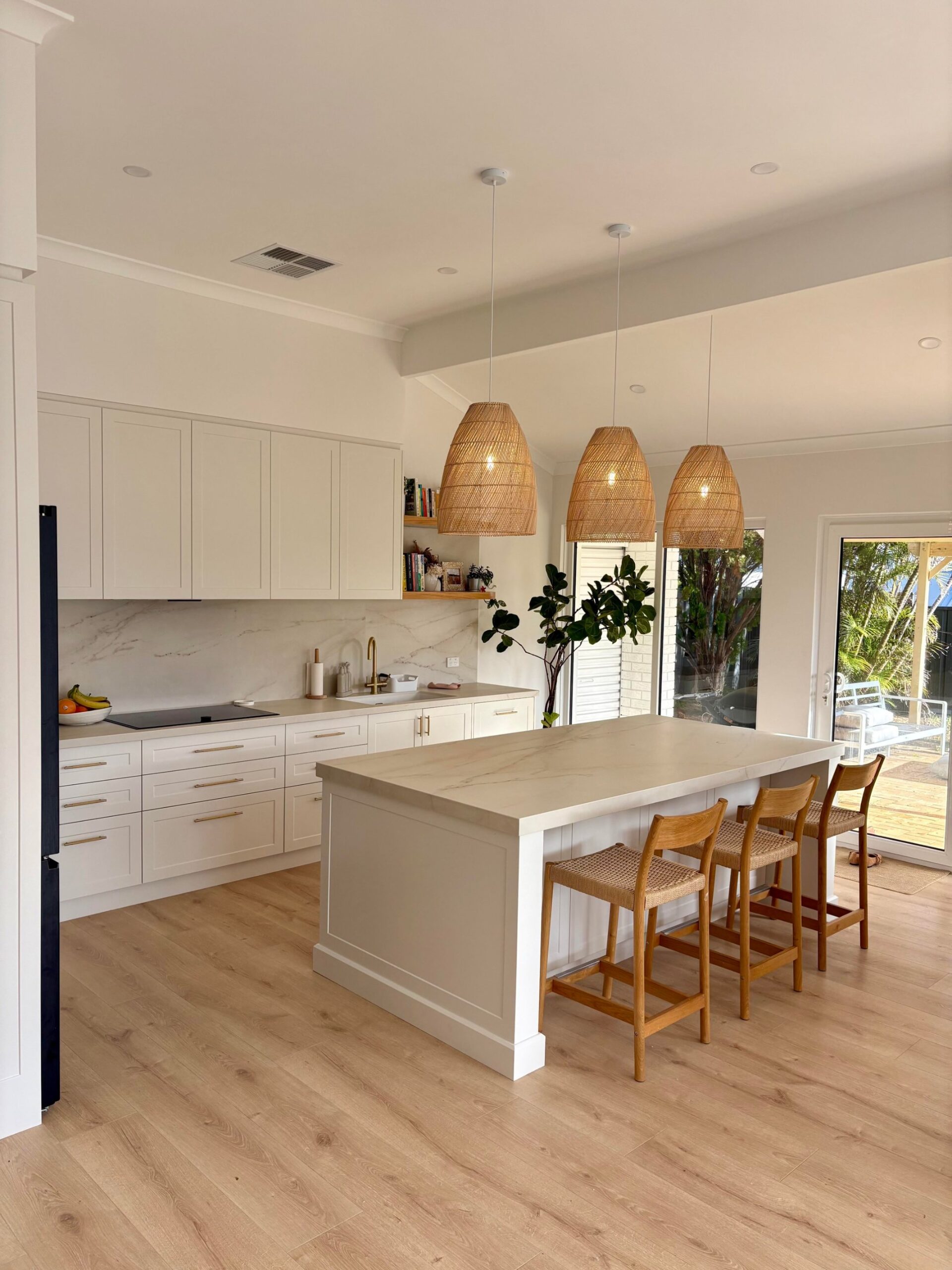 Bright white kitchen with a long Dekton island, three woven rattan pendant lights, brass tapware, and sliding doors opening to a leafy patio.