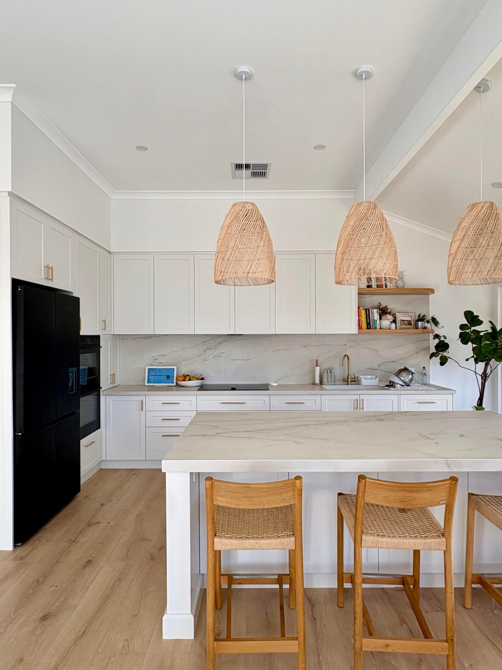 Wide shot of the kitchen showing the full L-shape, rattan pendants, three woven bar stools, black fridge, and a fiddle leaf fig by the back doors.