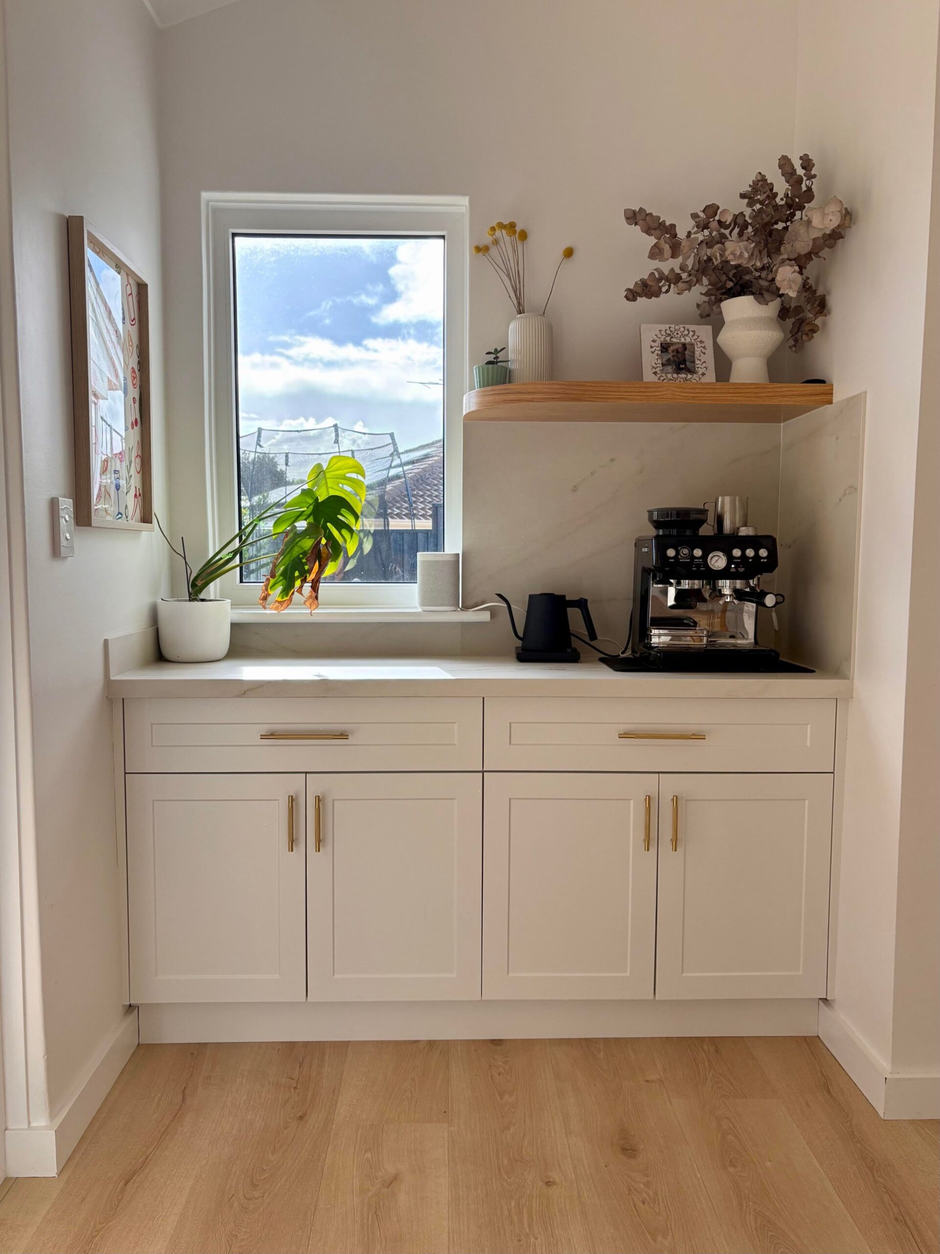 Small coffee station with white cabinets, brass pulls, a black espresso machine and kettle, a monstera plant on the window ledge, and marble backsplash.