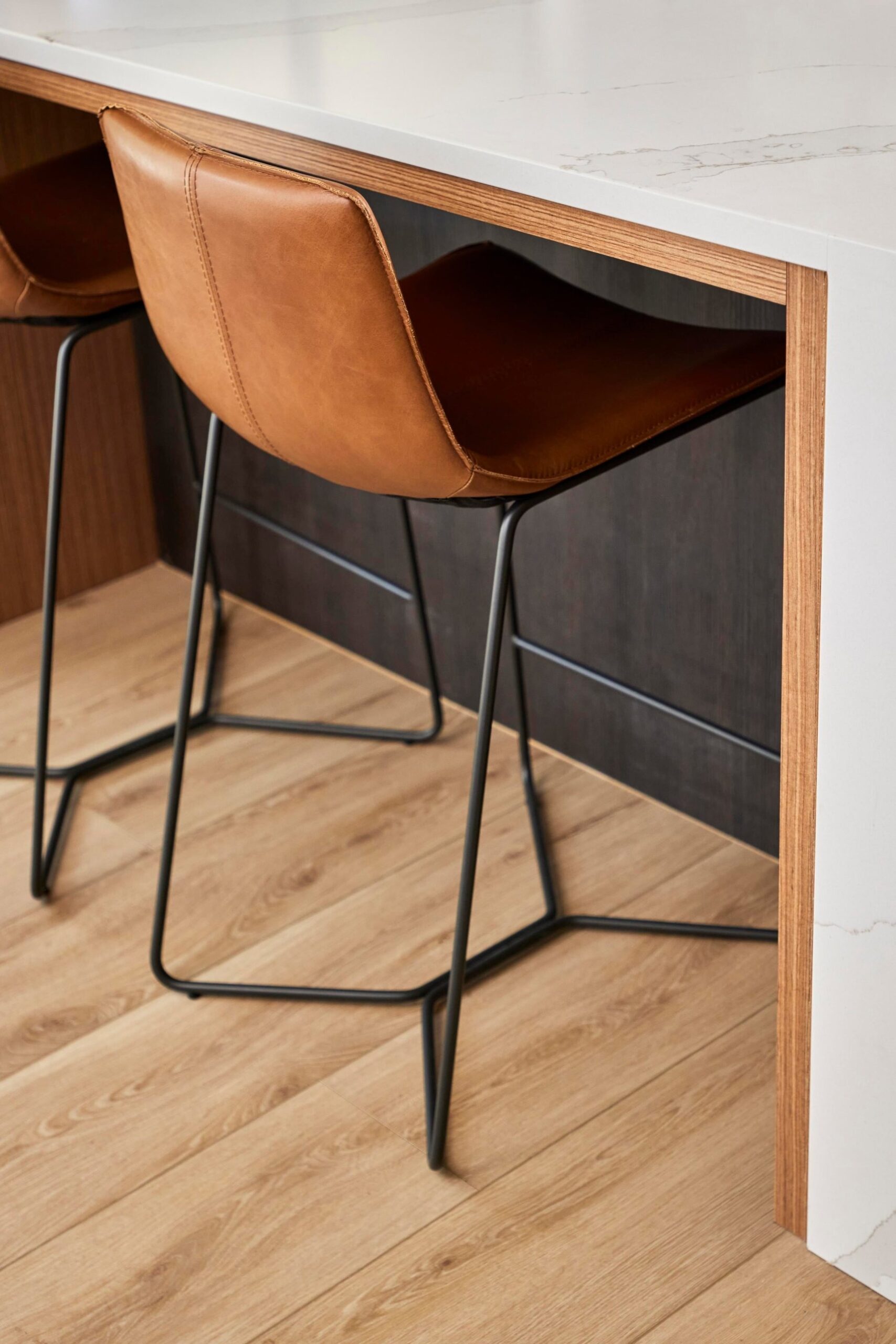 Close view of two cognac leather sled bar stools tucked under a white quartz waterfall island framed in wood veneer, over a light oak floor.